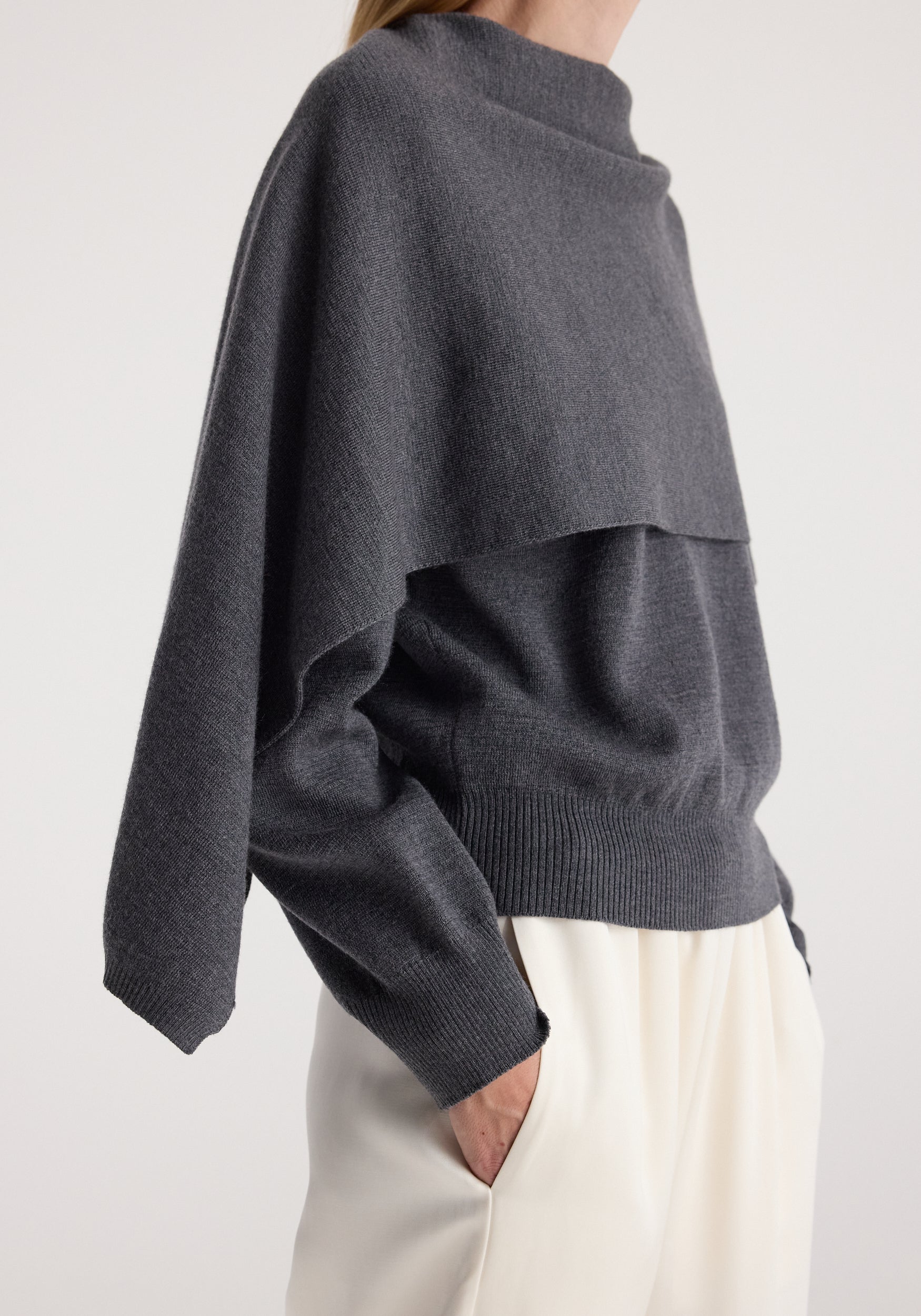Side view of a person wearing a dark grey melange knitted Cape jumper with a unique layered design over cream pants against a plain background.