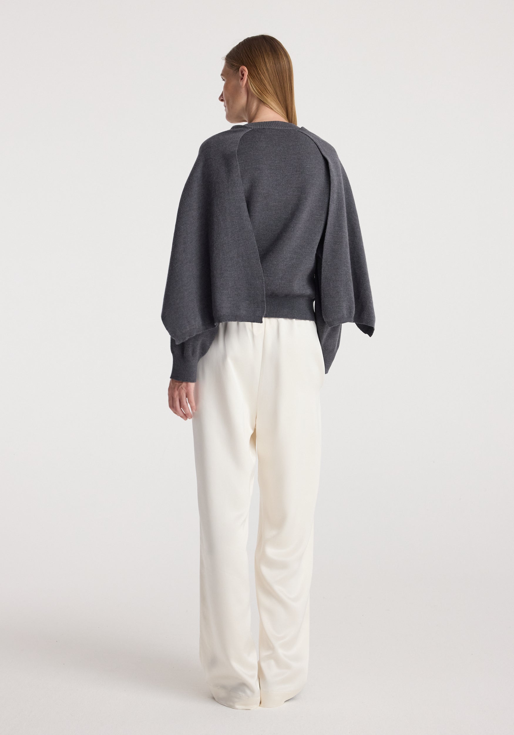 Woman wearing a dark grey melange cape jumper with long sleeves and a cape-like detail on the back, paired with loose white pants, standing against a plain background.