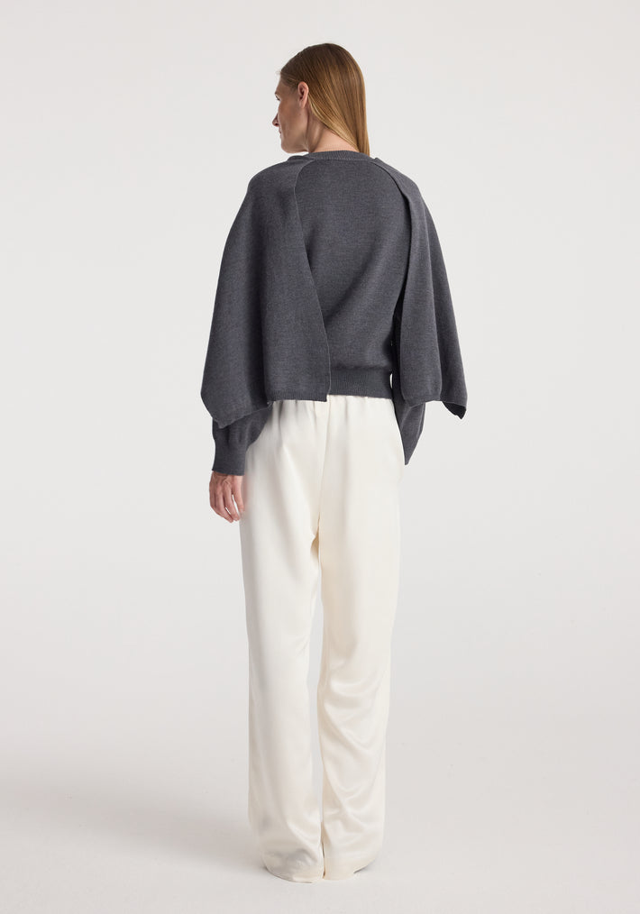 Woman wearing a dark grey melange cape jumper with long sleeves and a cape-like detail on the back, paired with loose white pants, standing against a plain background.