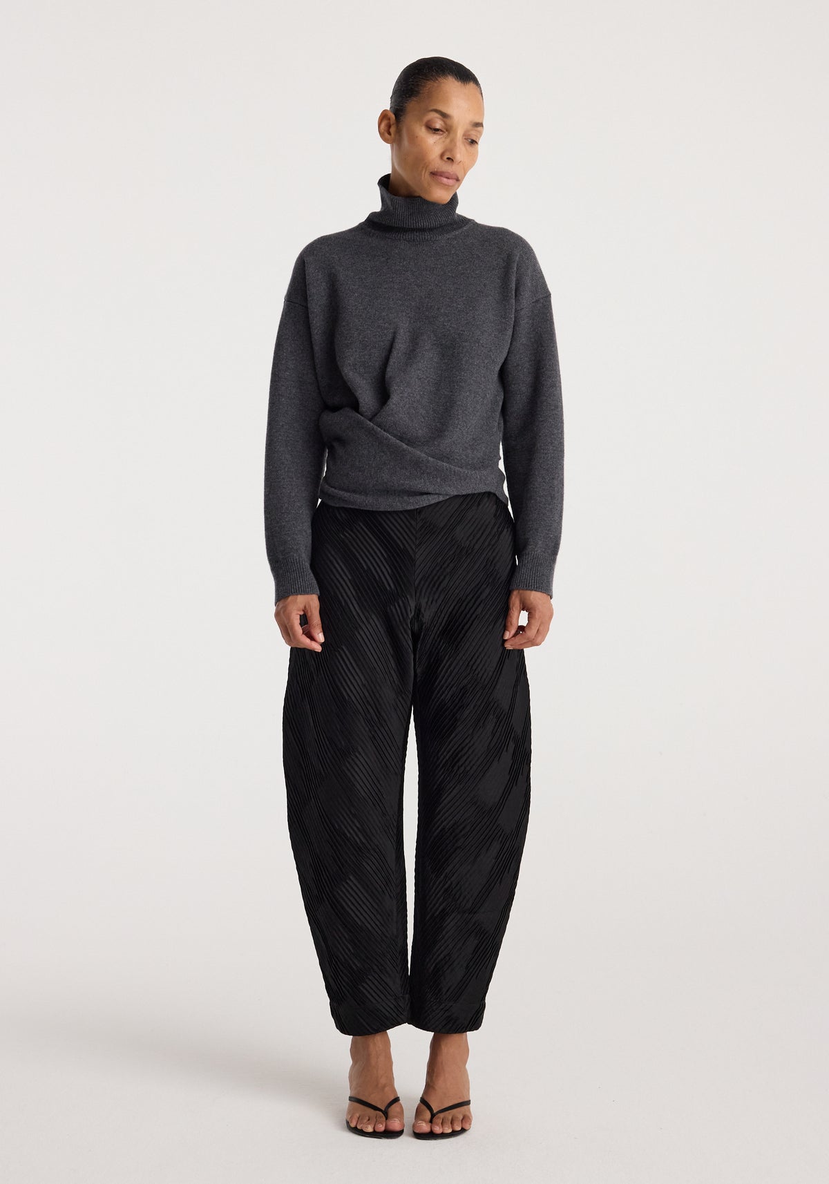 Woman wearing a Sculptural turtleneck | dark grey melange with long sleeves and a high collar, paired with black textured wide-leg pants and open-toe heels.