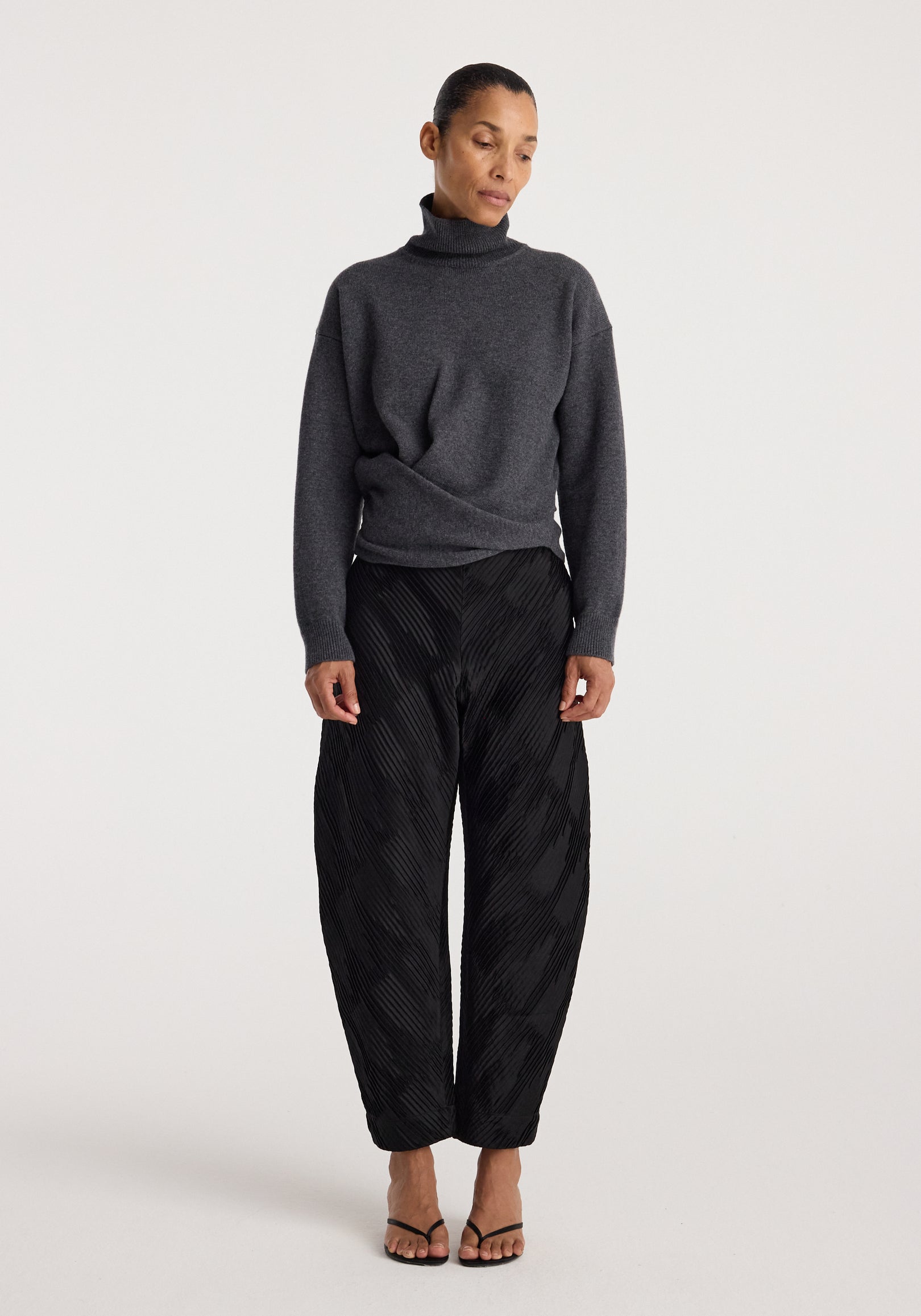 Woman wearing a Sculptural turtleneck | dark grey melange with long sleeves and a high collar, paired with black textured wide-leg pants and open-toe heels.