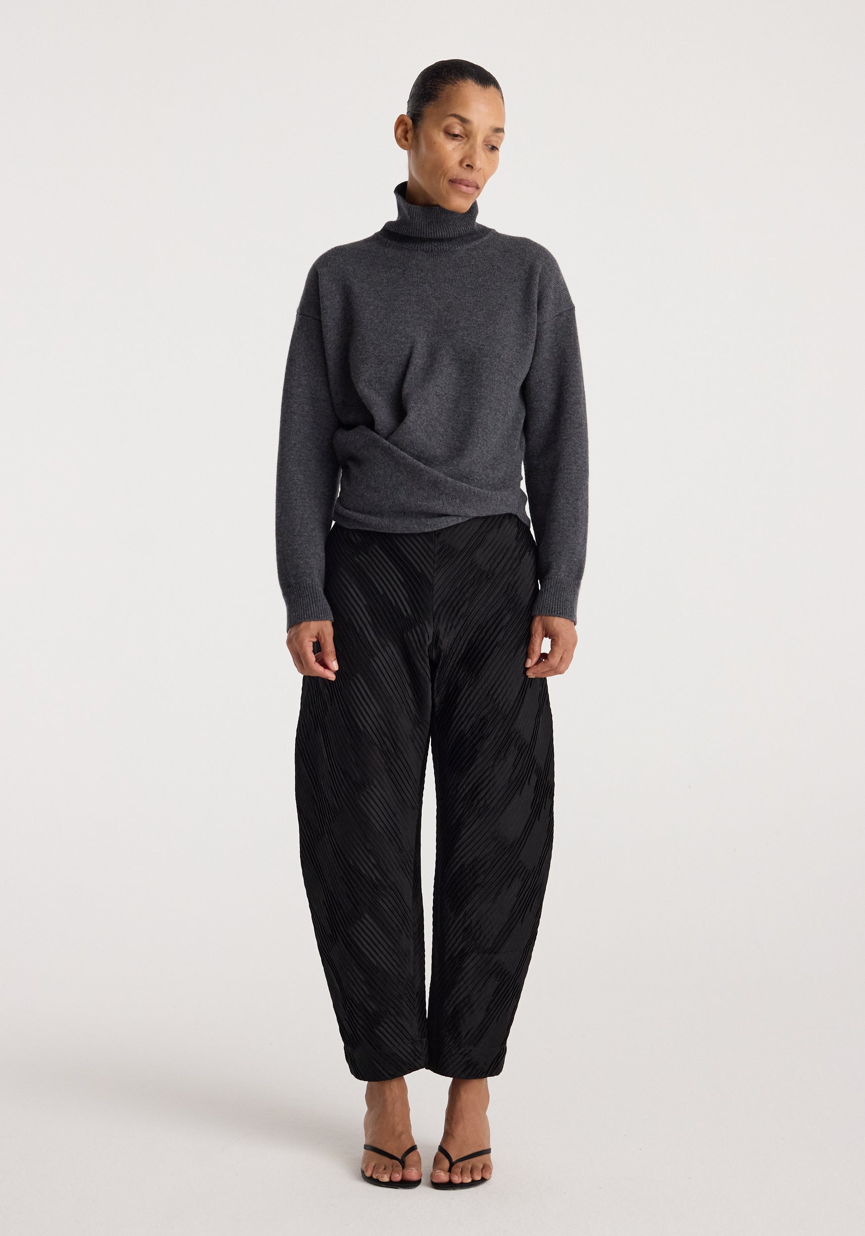 Woman wearing a Sculptural turtleneck | dark grey melange with long sleeves and a high collar, paired with black textured wide-leg pants and open-toe heels.
