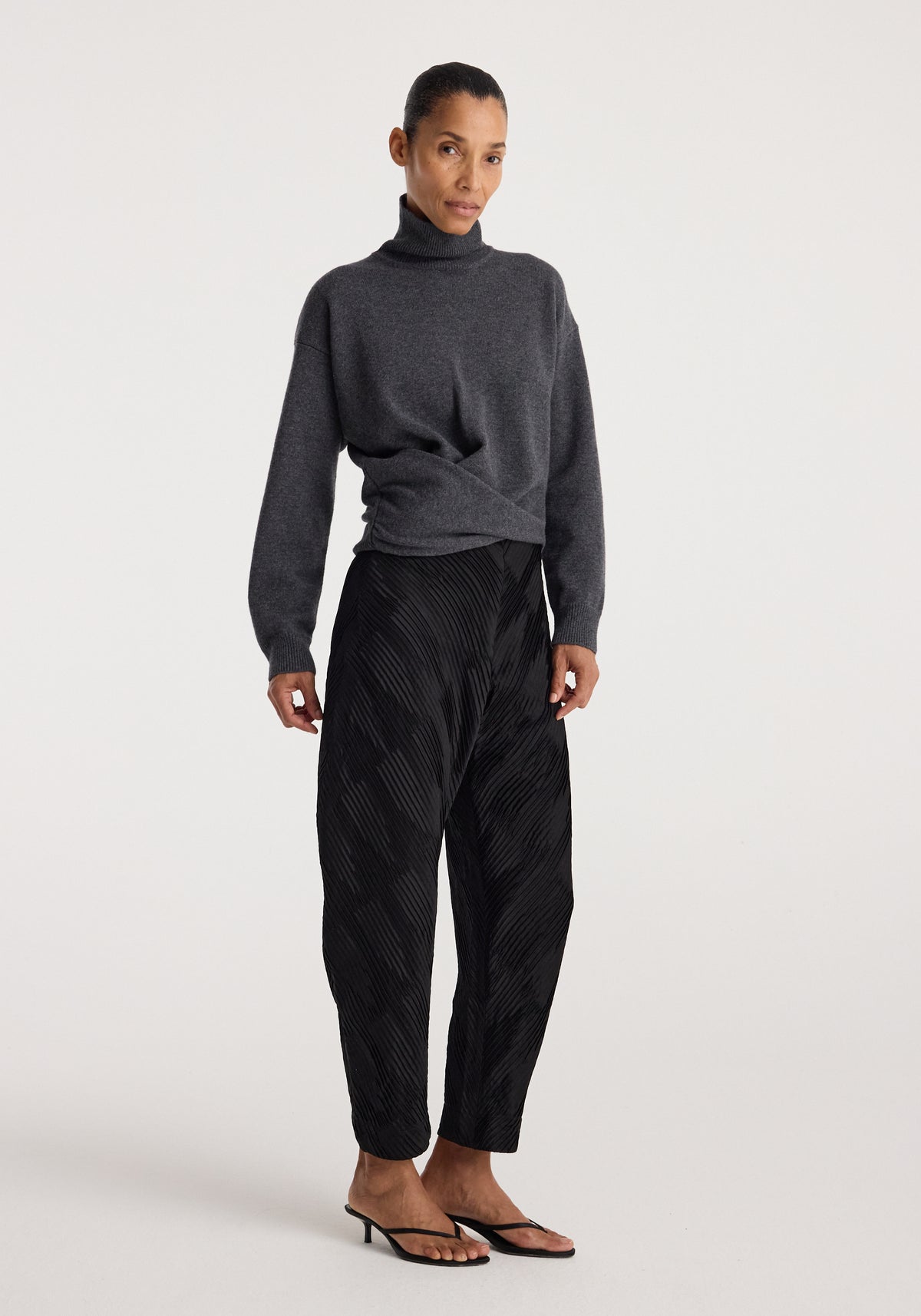 Woman wearing Sculptural turtleneck | dark grey melange paired with black patterned loose trousers and black heeled sandals standing against a plain light background.
