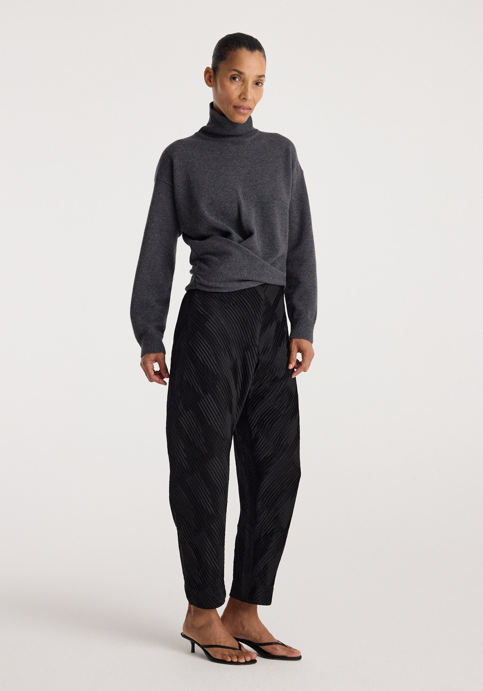 Woman wearing Sculptural turtleneck | dark grey melange paired with black patterned loose trousers and black heeled sandals standing against a plain light background.