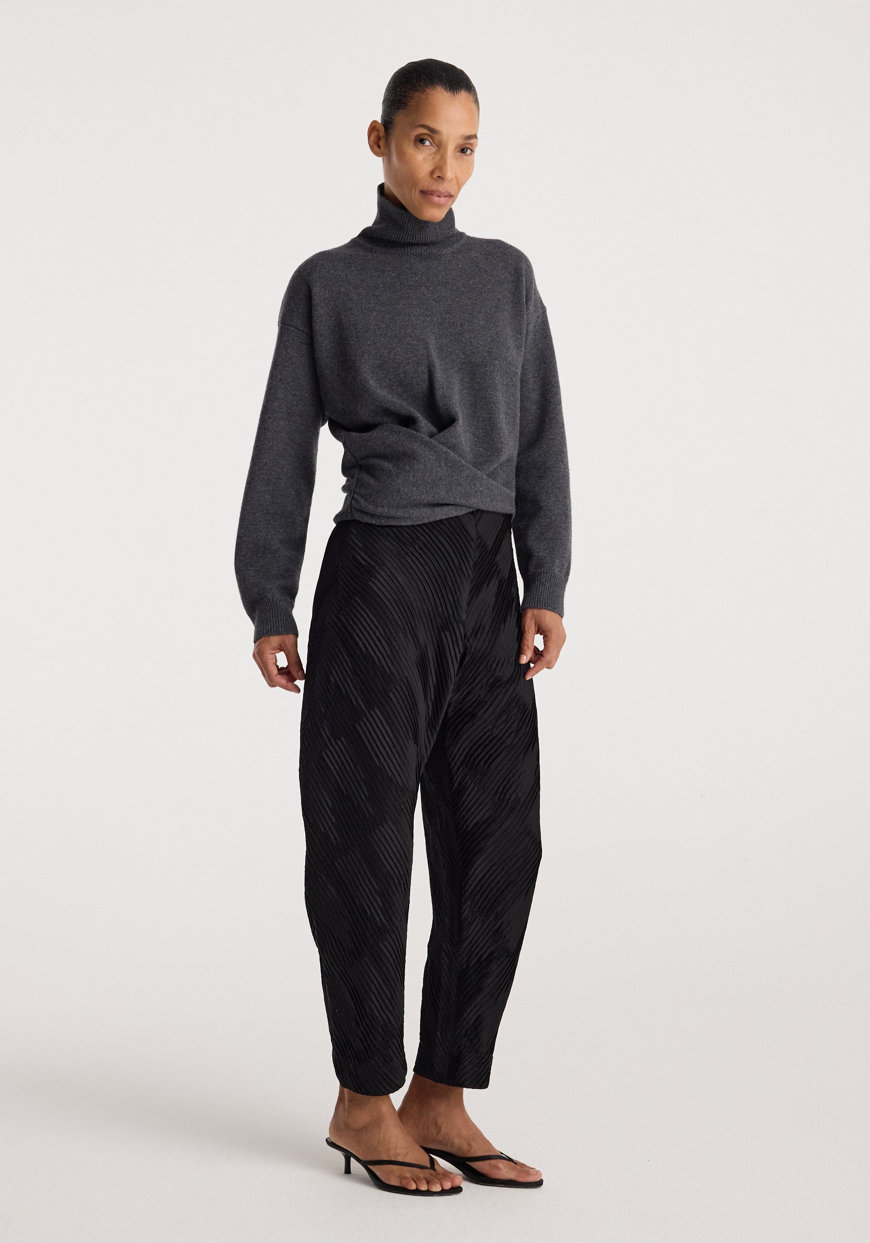 Woman wearing Sculptural turtleneck | dark grey melange paired with black patterned loose trousers and black heeled sandals standing against a plain light background.