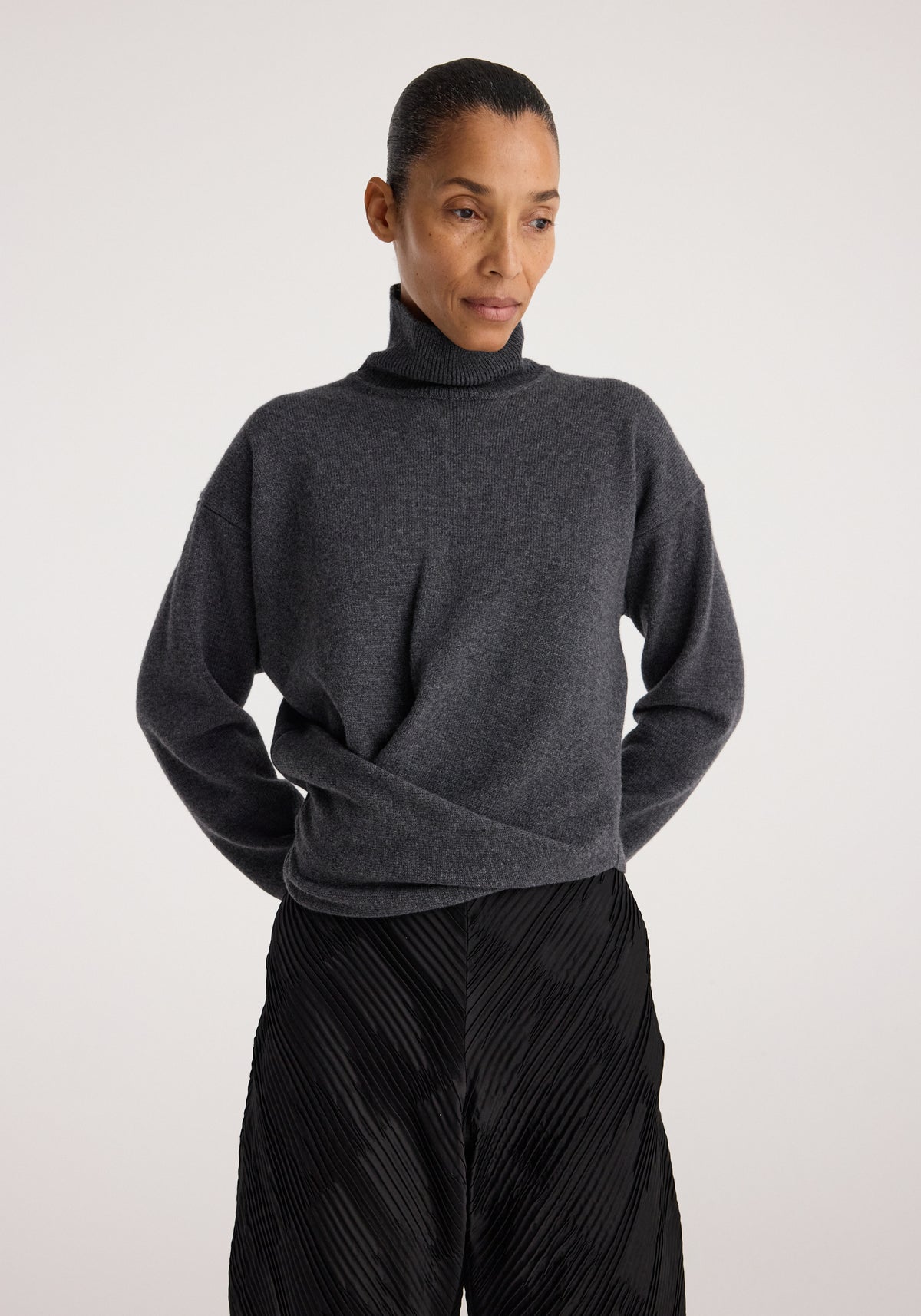 Woman wearing a Sculptural turtleneck | dark grey melange with a relaxed fit and long sleeves, paired with textured black pants against a plain background.