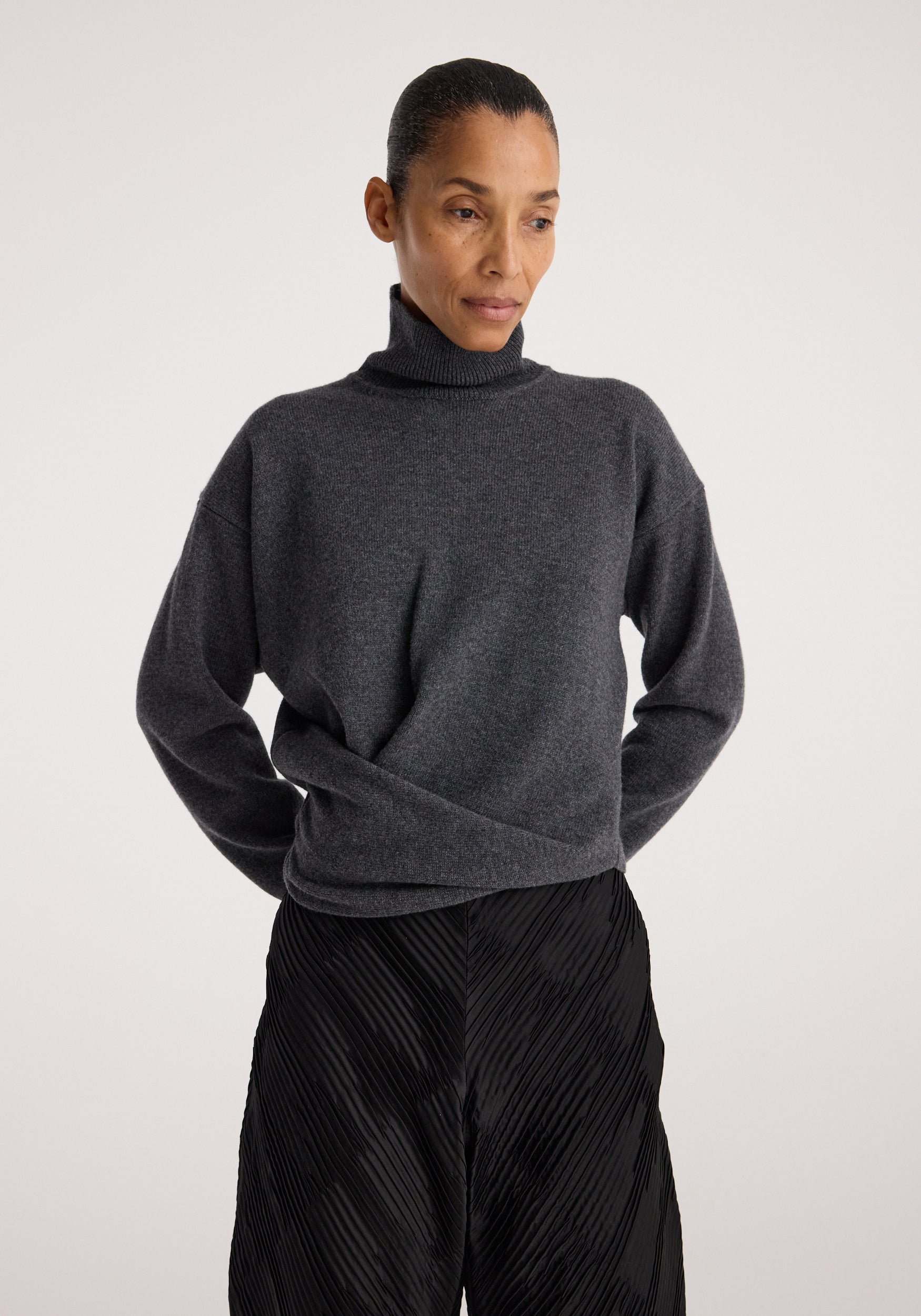 Woman wearing a Sculptural turtleneck | dark grey melange with a relaxed fit and long sleeves, paired with textured black pants against a plain background.