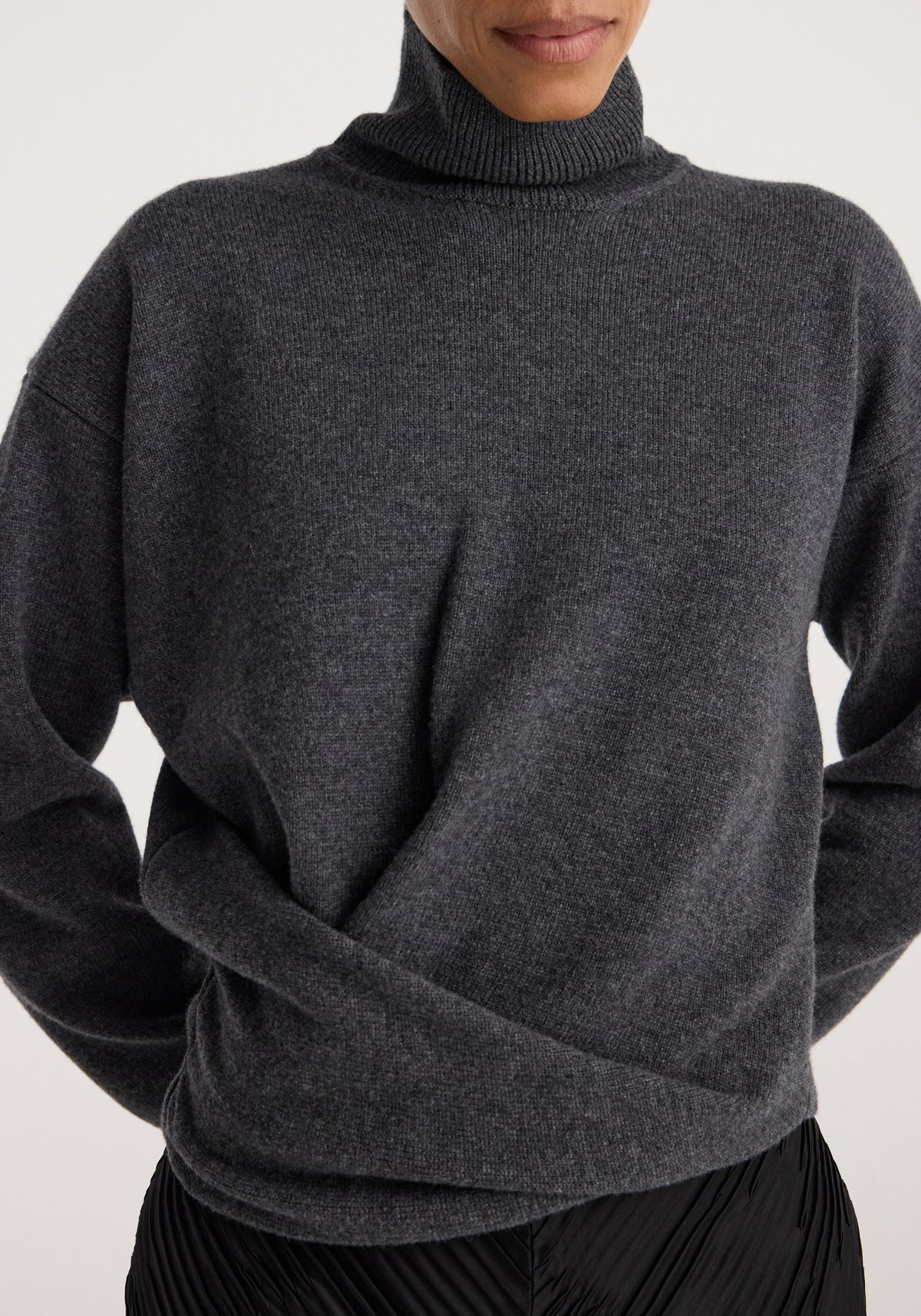 Close-up of a person wearing a soft Sculptural turtleneck | dark grey melange with a high neck and relaxed fit, paired with black pleated bottoms.