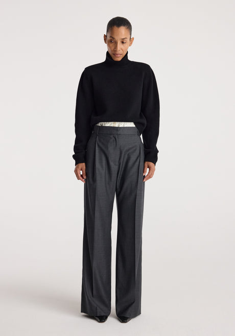 Woman wearing a black Statement shoulder turtleneck | black with long sleeves, styled with high-waisted gray wide-leg trousers, standing against a plain background.