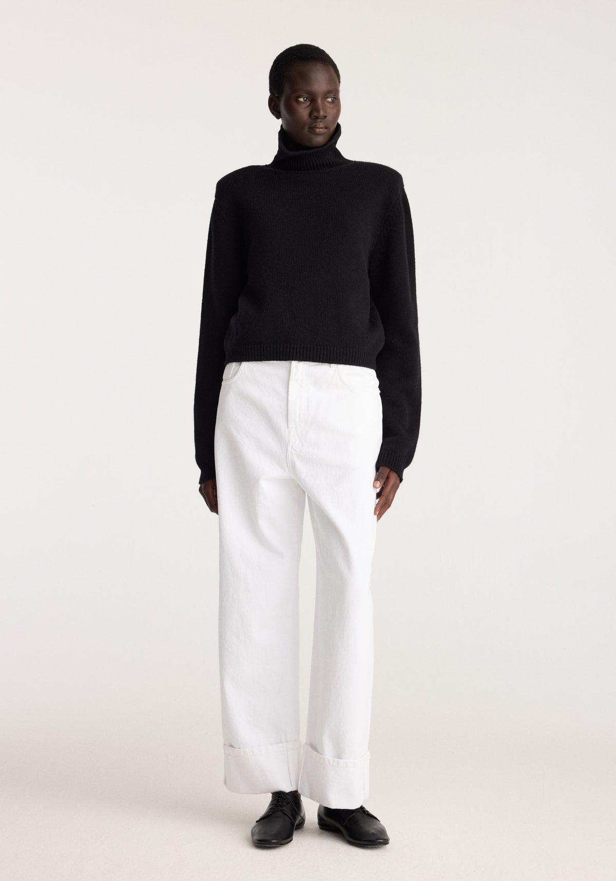 Statement shoulder turtleneck | black modeled with white cuffed trousers and black shoes in a minimalist studio setting