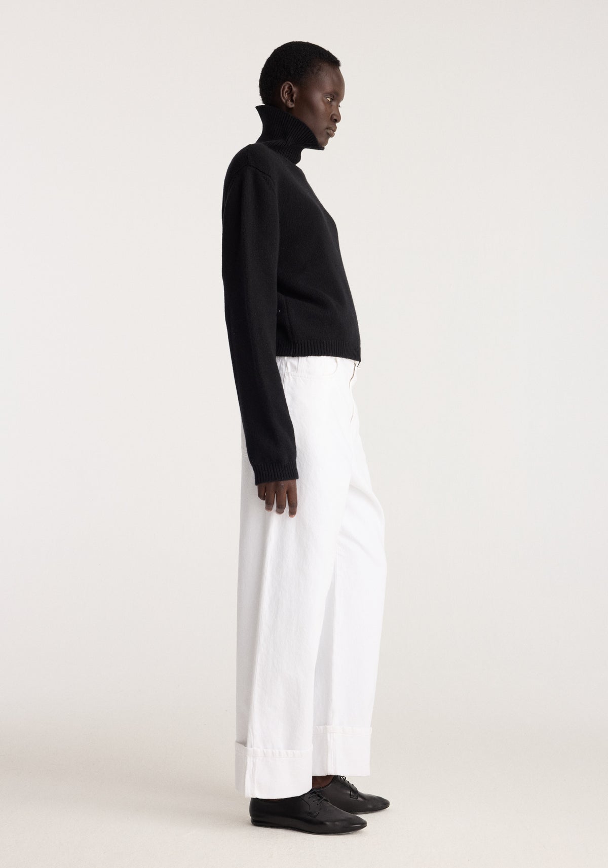 Statement shoulder turtleneck | black worn with white wide leg trousers and black shoes in a side profile studio portrait