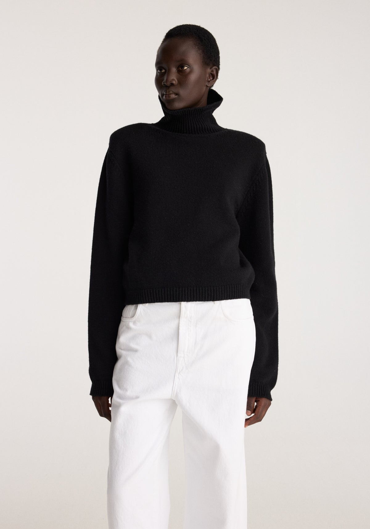 Model wearing Statement shoulder turtleneck | black with a high neck and long sleeves styled with relaxed white trousers against a neutral background