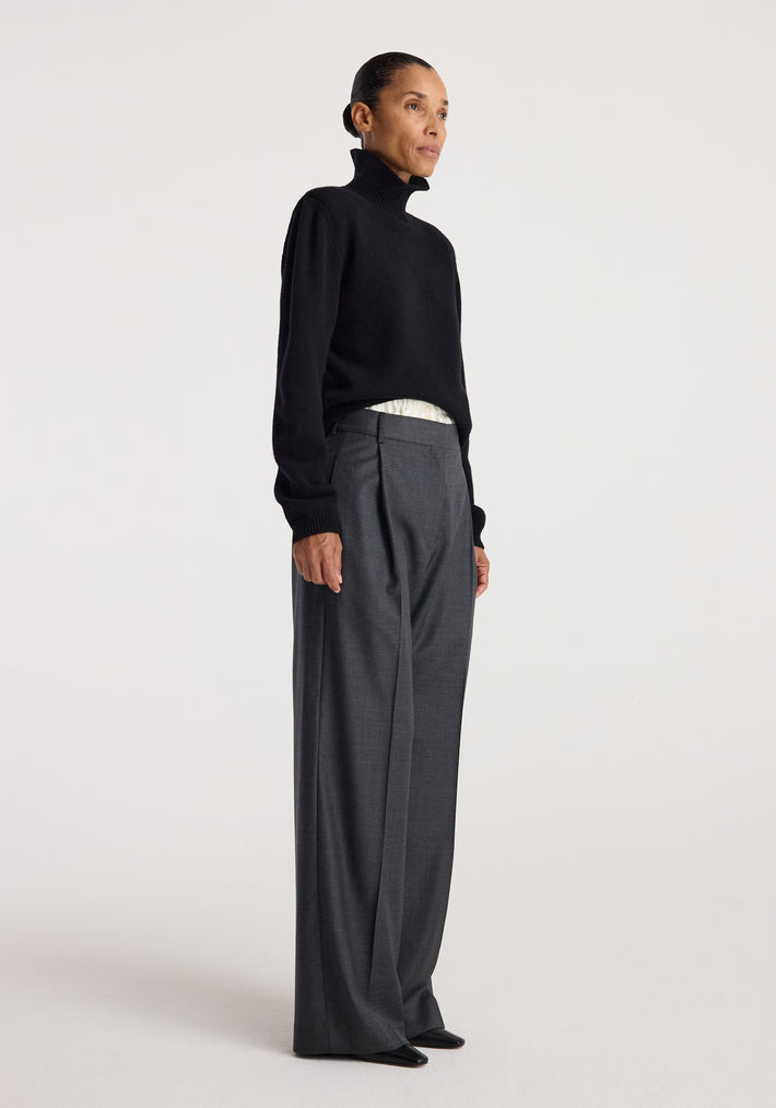 Woman wearing a black Statement shoulder turtleneck | black with long sleeves and a high collar, paired with gray wide-leg trousers and black shoes, standing against a light background.