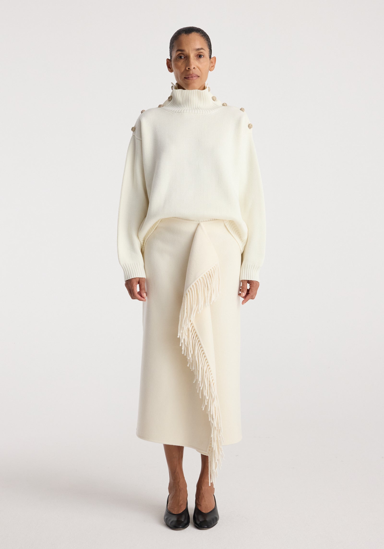 Woman wearing an elegant cream outfit featuring an Uneven sailor polo | cream with button details on the shoulders and a long skirt with a fringed asymmetrical overlay, paired with black shoes.