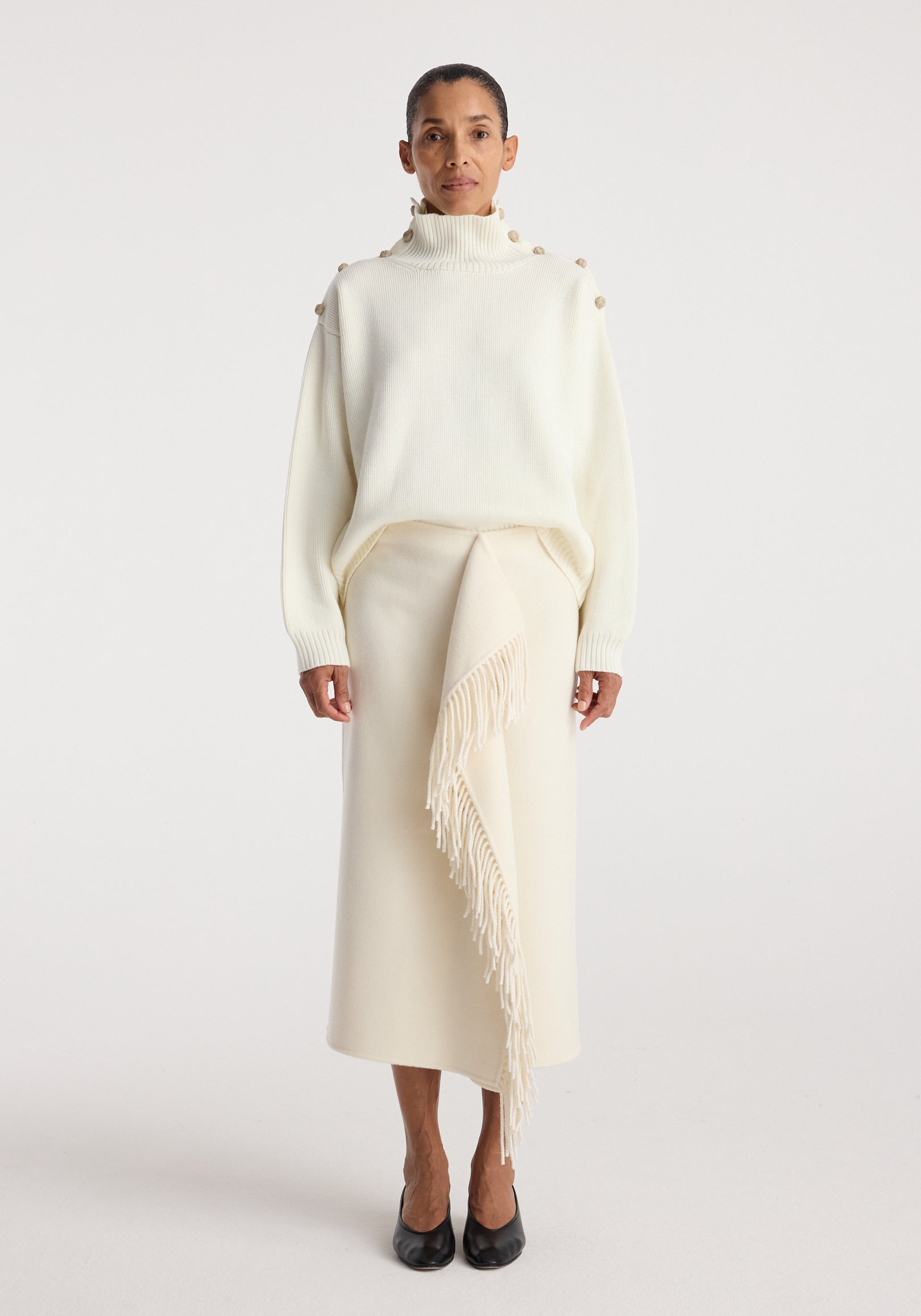 Woman wearing an elegant cream outfit featuring an Uneven sailor polo | cream with button details on the shoulders and a long skirt with a fringed asymmetrical overlay, paired with black shoes.