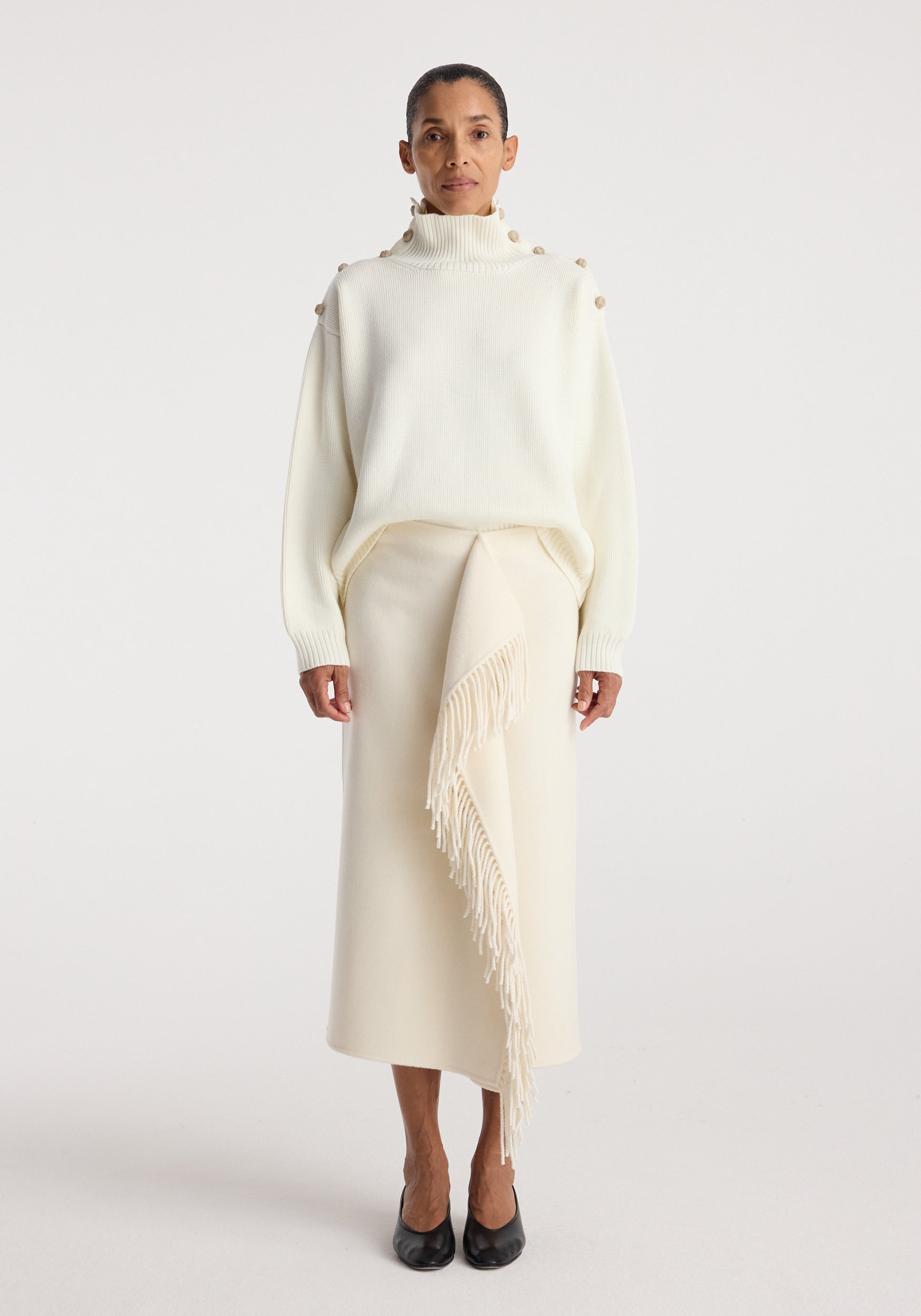 Woman wearing an elegant cream outfit featuring an Uneven sailor polo | cream with button details on the shoulders and a long skirt with a fringed asymmetrical overlay, paired with black shoes.