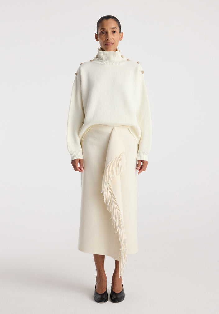 Woman wearing an elegant cream outfit featuring an Uneven sailor polo | cream with button details on the shoulders and a long skirt with a fringed asymmetrical overlay, paired with black shoes.