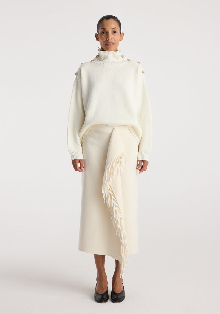 Woman wearing an elegant cream outfit featuring an Uneven sailor polo | cream with button details on the shoulders and a long skirt with a fringed asymmetrical overlay, paired with black shoes.