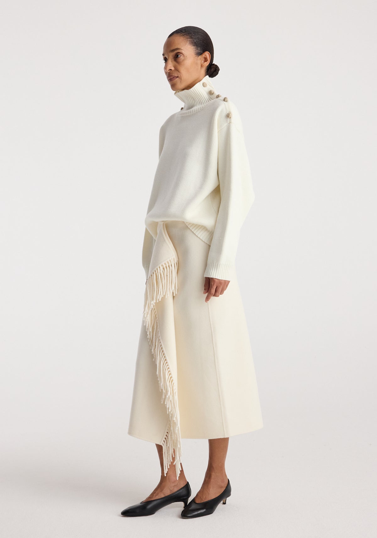 Woman wearing an elegant cream-colored knit turtleneck with button details on the shoulder and a matching cream midi skirt with fringed accents, styled with black low-heeled shoes, featuring the Uneven sailor polo | cream.
