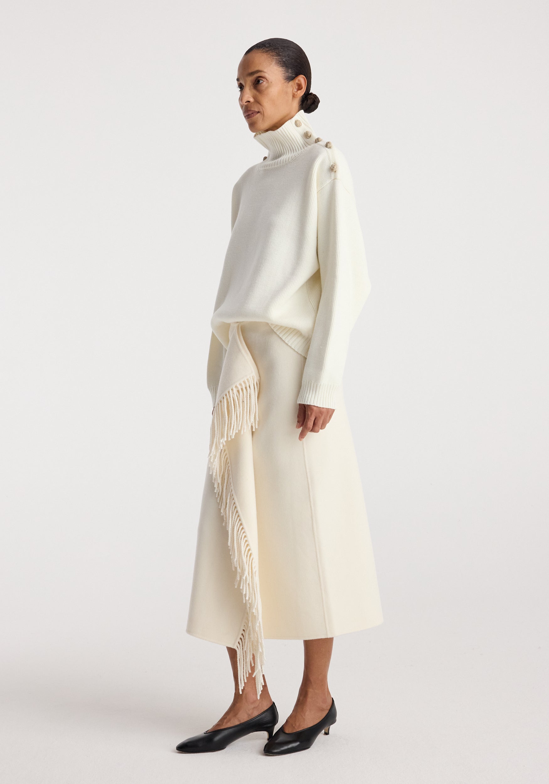 Woman wearing an elegant cream-colored knit turtleneck with button details on the shoulder and a matching cream midi skirt with fringed accents, styled with black low-heeled shoes, featuring the Uneven sailor polo | cream.