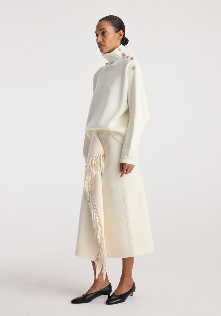 Woman wearing an elegant cream outfit featuring an Uneven sailor polo | cream with button details on the shoulders and a long skirt with a fringed asymmetrical overlay, paired with black shoes.