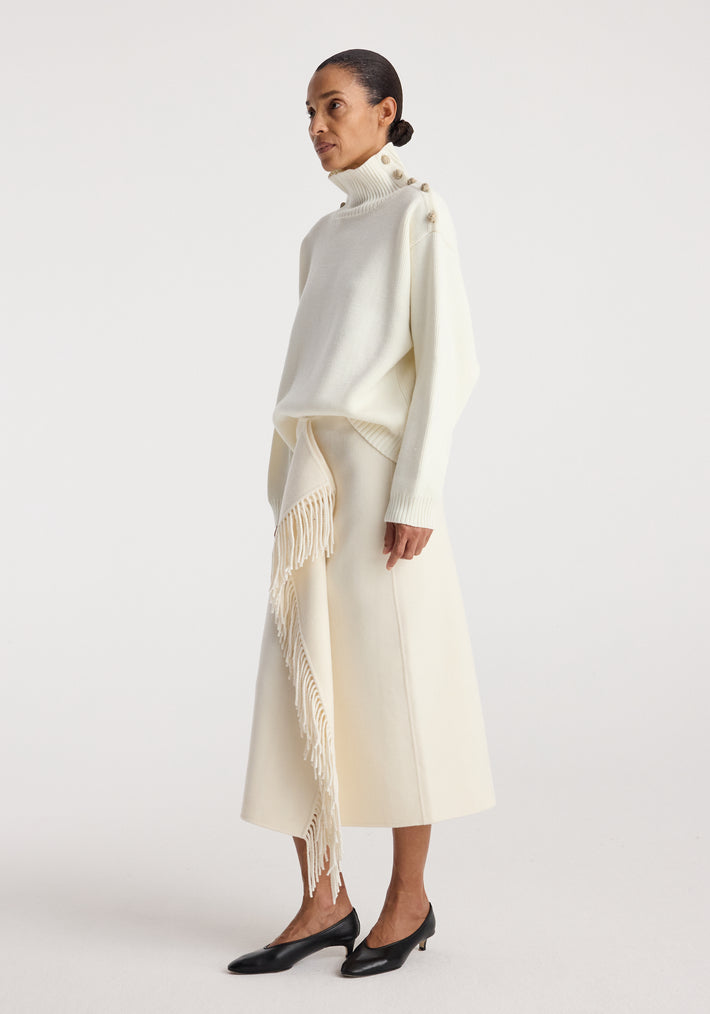 Woman wearing an elegant cream-colored knit turtleneck with button details on the shoulder and a matching cream midi skirt with fringed accents, styled with black low-heeled shoes, featuring the Uneven sailor polo | cream.