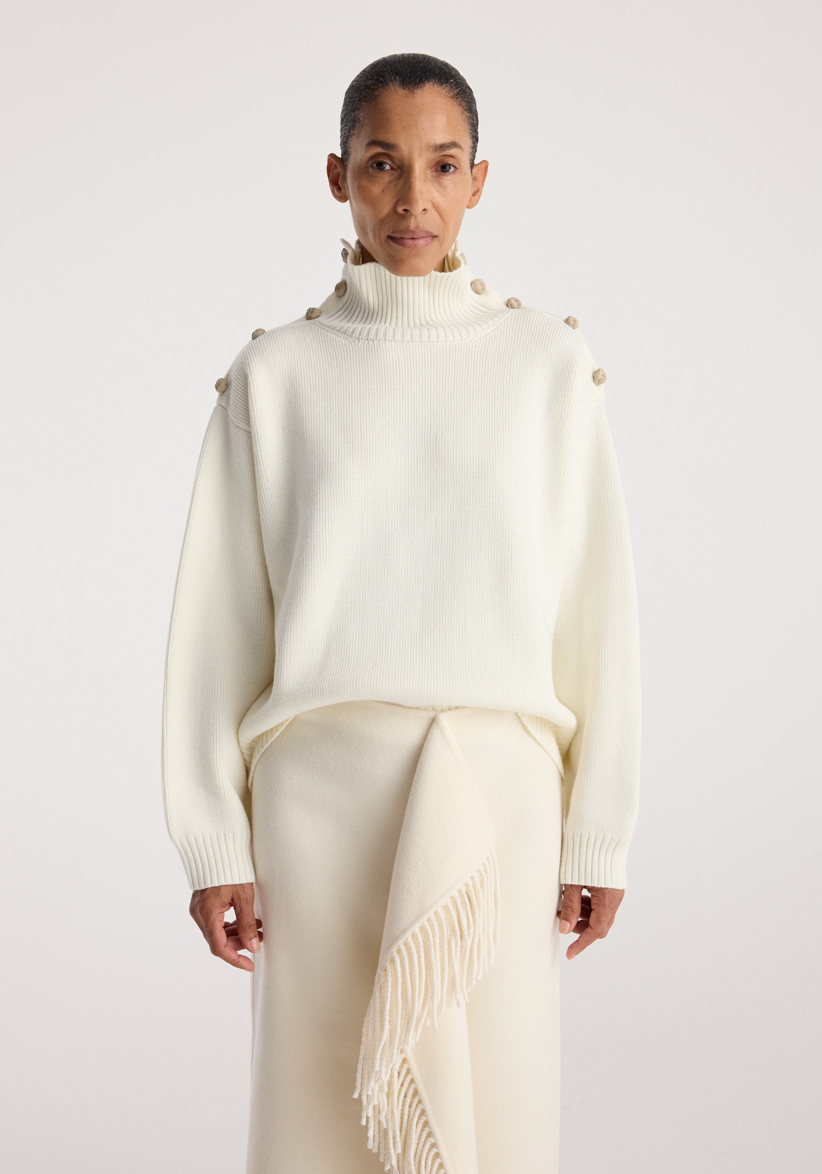 Woman wearing a cream high-neck sweater with buttons along the shoulders and upper sleeves, paired with a cream skirt featuring a fringed detail, styled as Uneven sailor polo | cream.