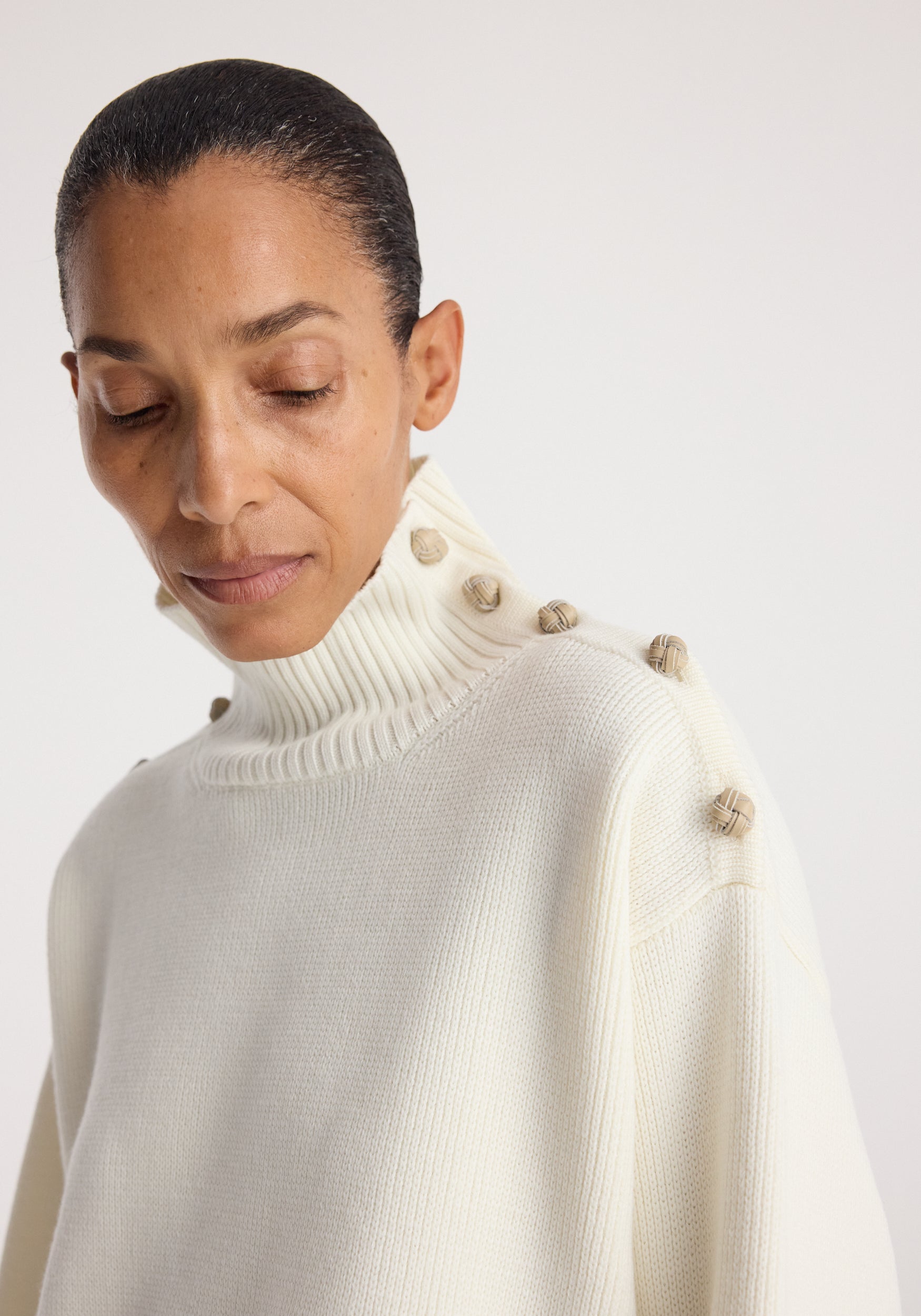Close-up of a woman wearing an uneven sailor polo | cream with a high ribbed collar and decorative buttons along the shoulder and collar.