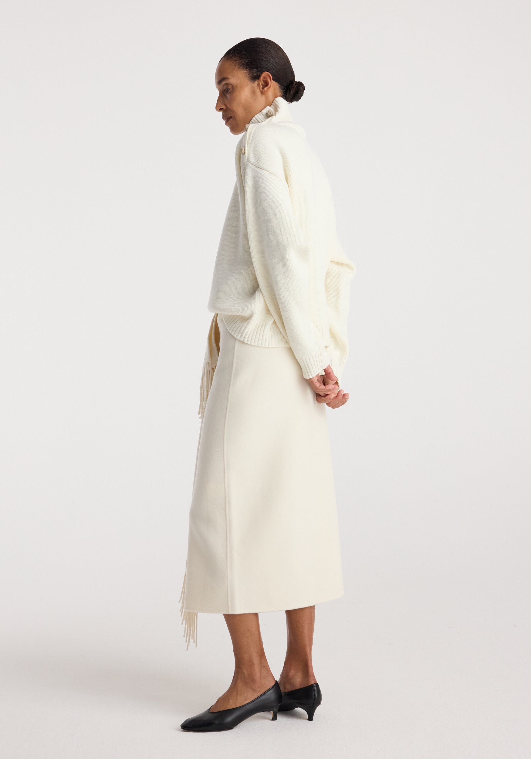 Side view of a woman wearing a cream-colored Uneven sailor polo | cream with a midi skirt and black low-heeled shoes against a plain white background.