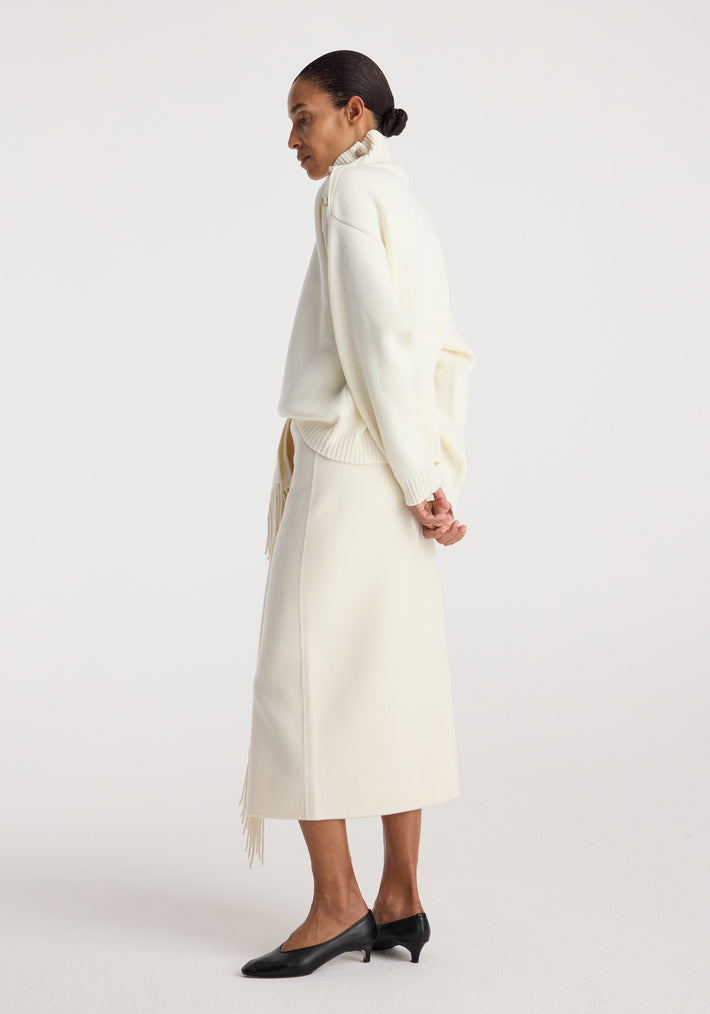 Side view of a woman wearing a cream-colored Uneven sailor polo | cream with a midi skirt and black low-heeled shoes against a plain white background.