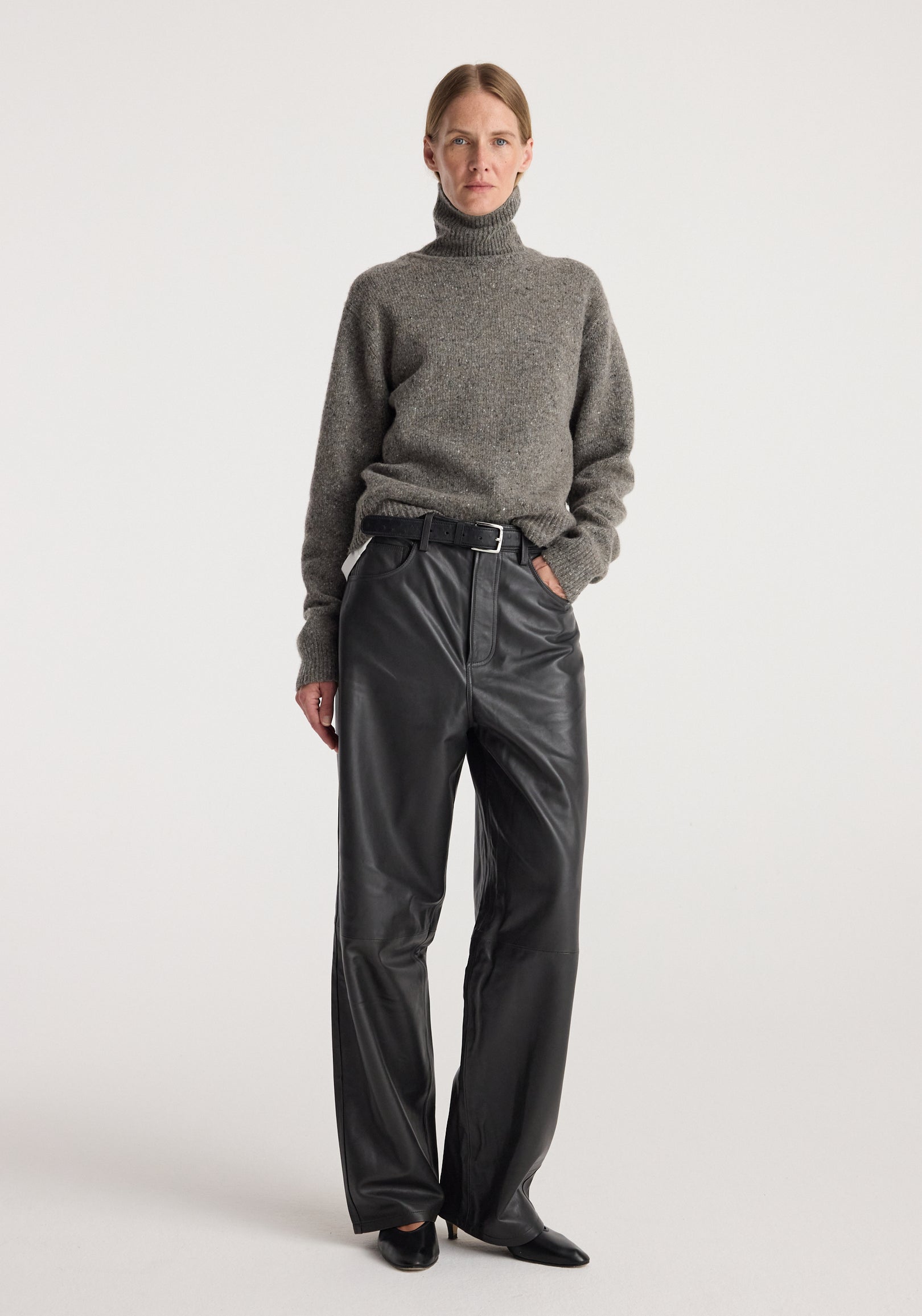 Woman wearing a grey mouliné textured knit Mouliné turtleneck | grey mouliné paired with black leather wide-leg pants and black pointed shoes standing against a plain background.