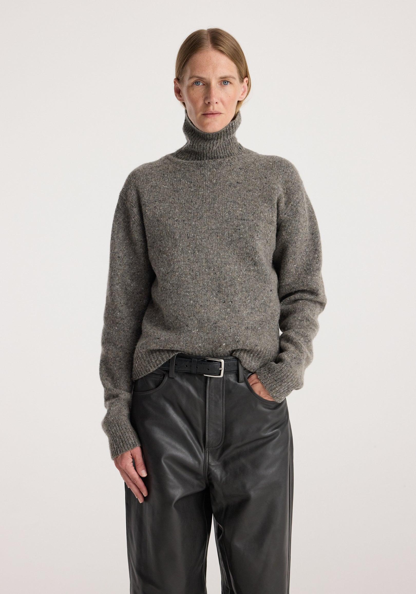 A woman wearing a cosy grey mouliné high-neck sweater with long sleeves paired with black leather pants and a black belt, styled in a minimalistic setting, featuring the Mouliné turtleneck | grey mouliné.