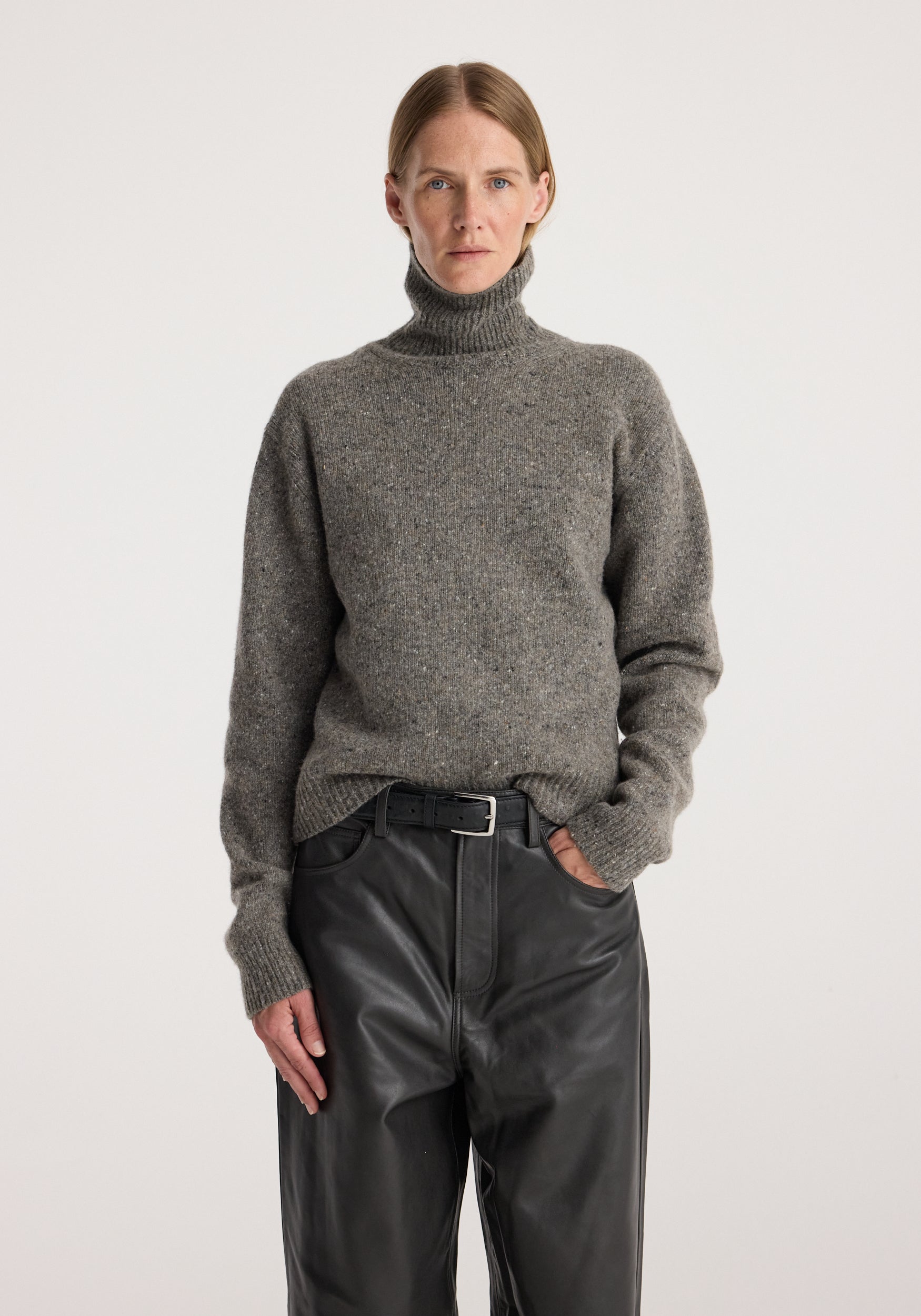 A woman wearing a cosy grey mouliné high-neck sweater with long sleeves paired with black leather pants and a black belt, styled in a minimalistic setting, featuring the Mouliné turtleneck | grey mouliné.