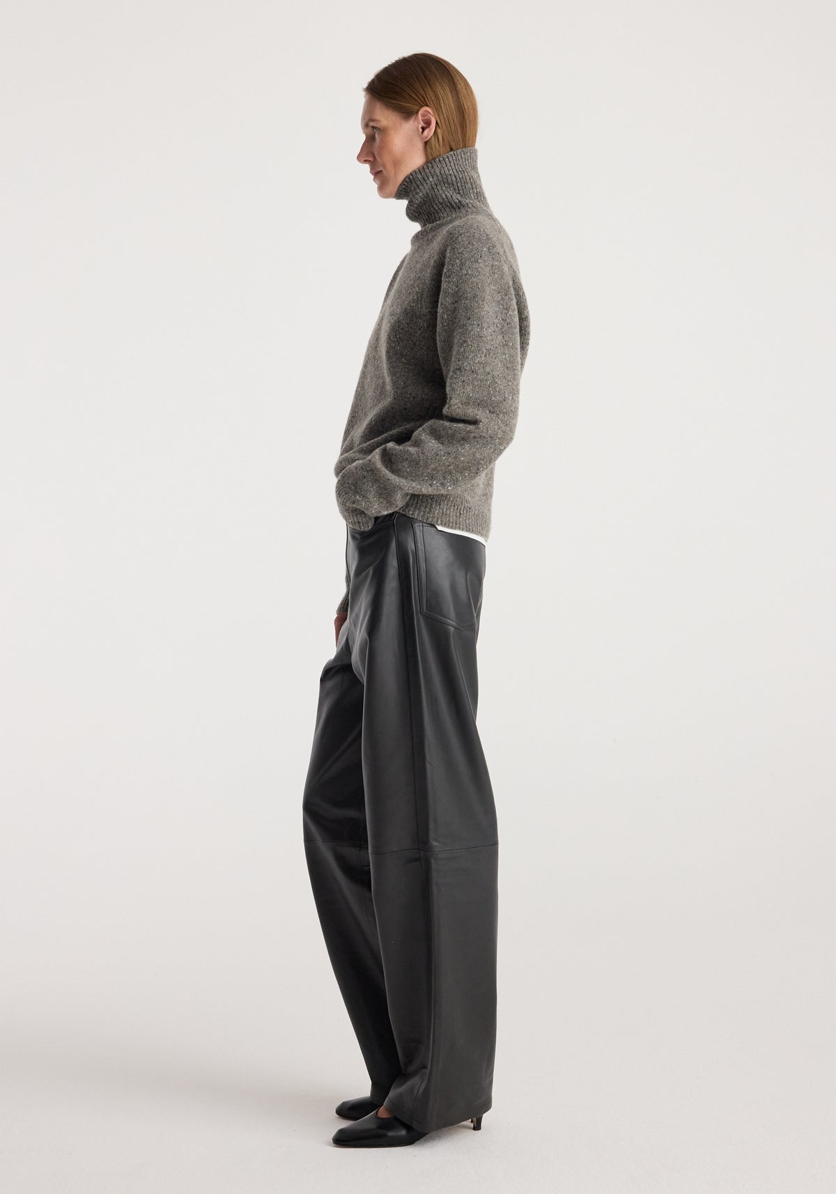 Woman wearing a grey textured Mouliné turtleneck | grey mouliné sweater paired with wide-leg black leather pants and black pointed-toe shoes, standing in profile against a plain background.