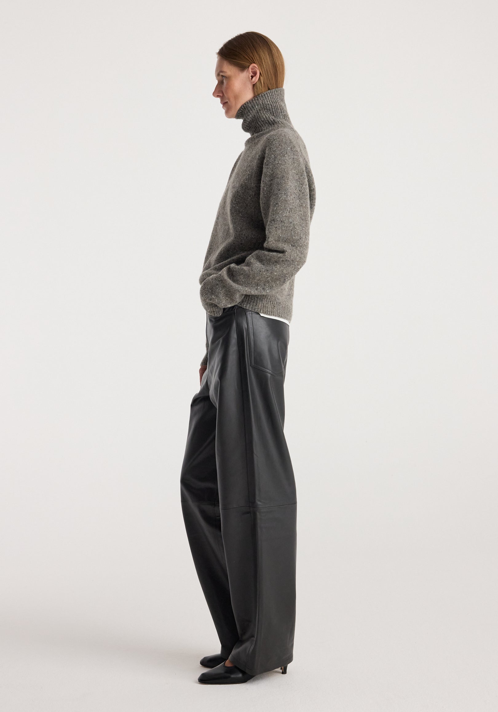 Woman wearing a grey textured Mouliné turtleneck | grey mouliné sweater paired with wide-leg black leather pants and black pointed-toe shoes, standing in profile against a plain background.