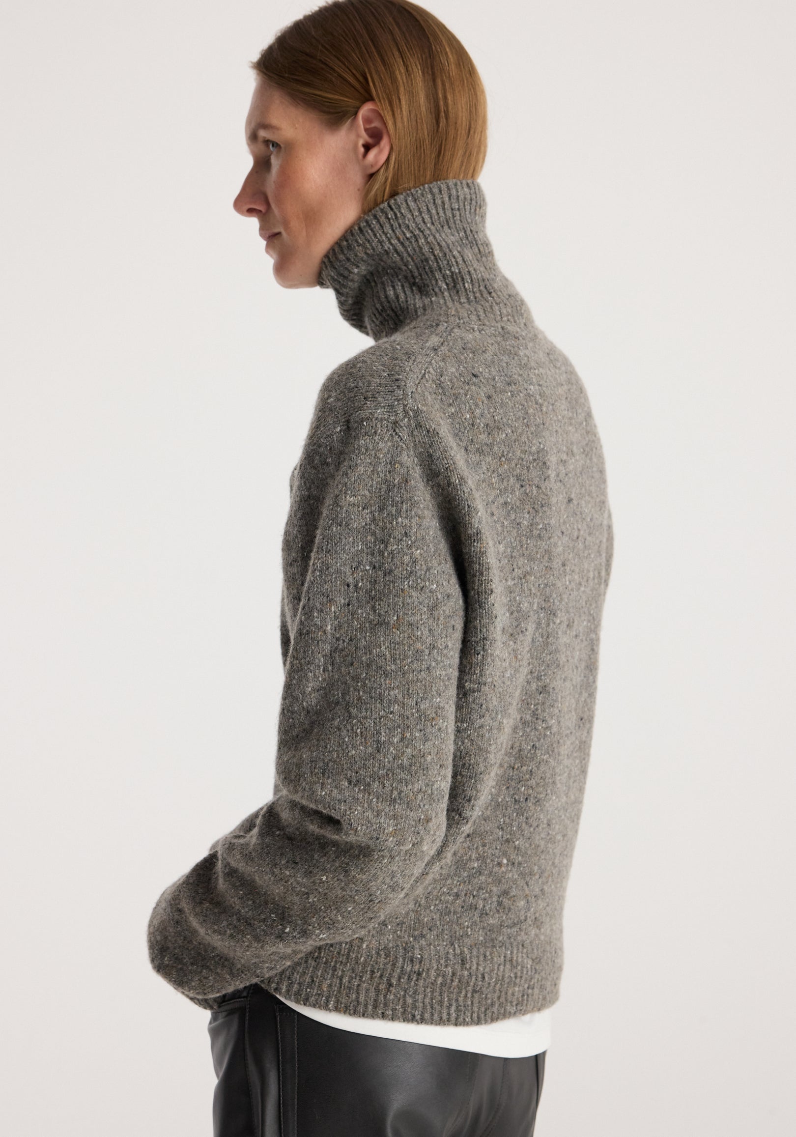 Side view of a person wearing a thick knit grey turtleneck sweater with a ribbed collar and hem, paired with black pants and a white shirt underneath, featuring the Mouliné turtleneck | grey mouliné.