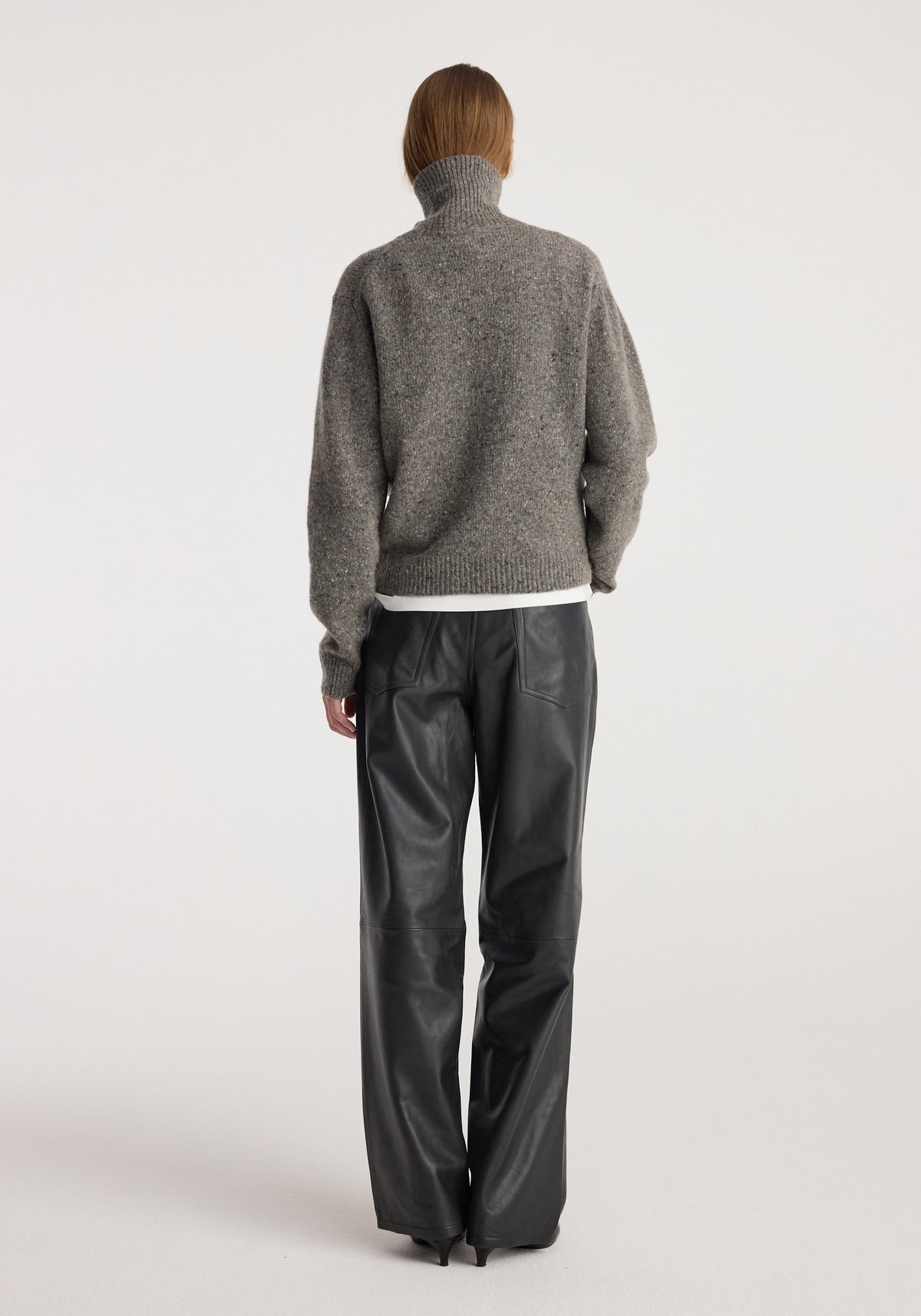 Rear view of a person wearing a grey Mouliné turtleneck | grey mouliné sweater with a high neck and long sleeves paired with black wide-leg leather pants against a plain white background.