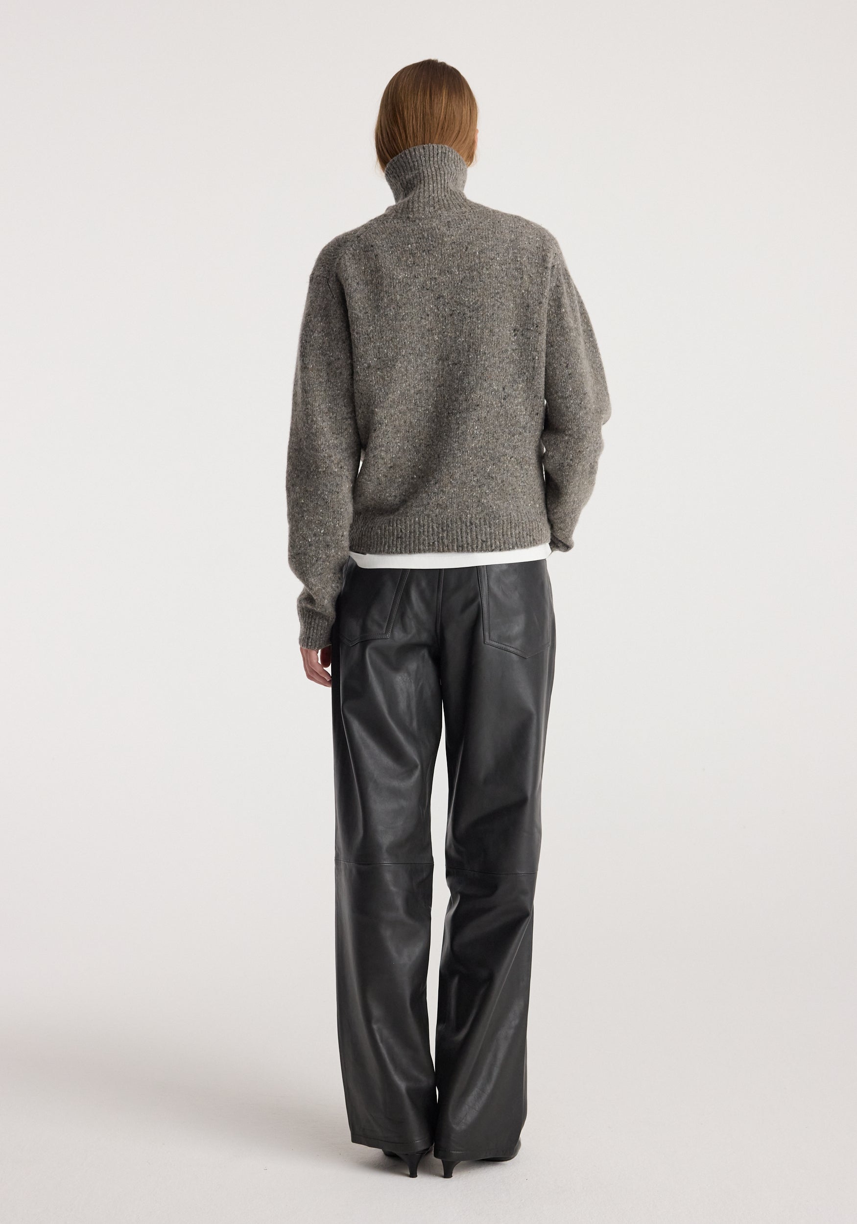 Rear view of a person wearing a grey Mouliné turtleneck | grey mouliné sweater with a high neck and long sleeves paired with black wide-leg leather pants against a plain white background.