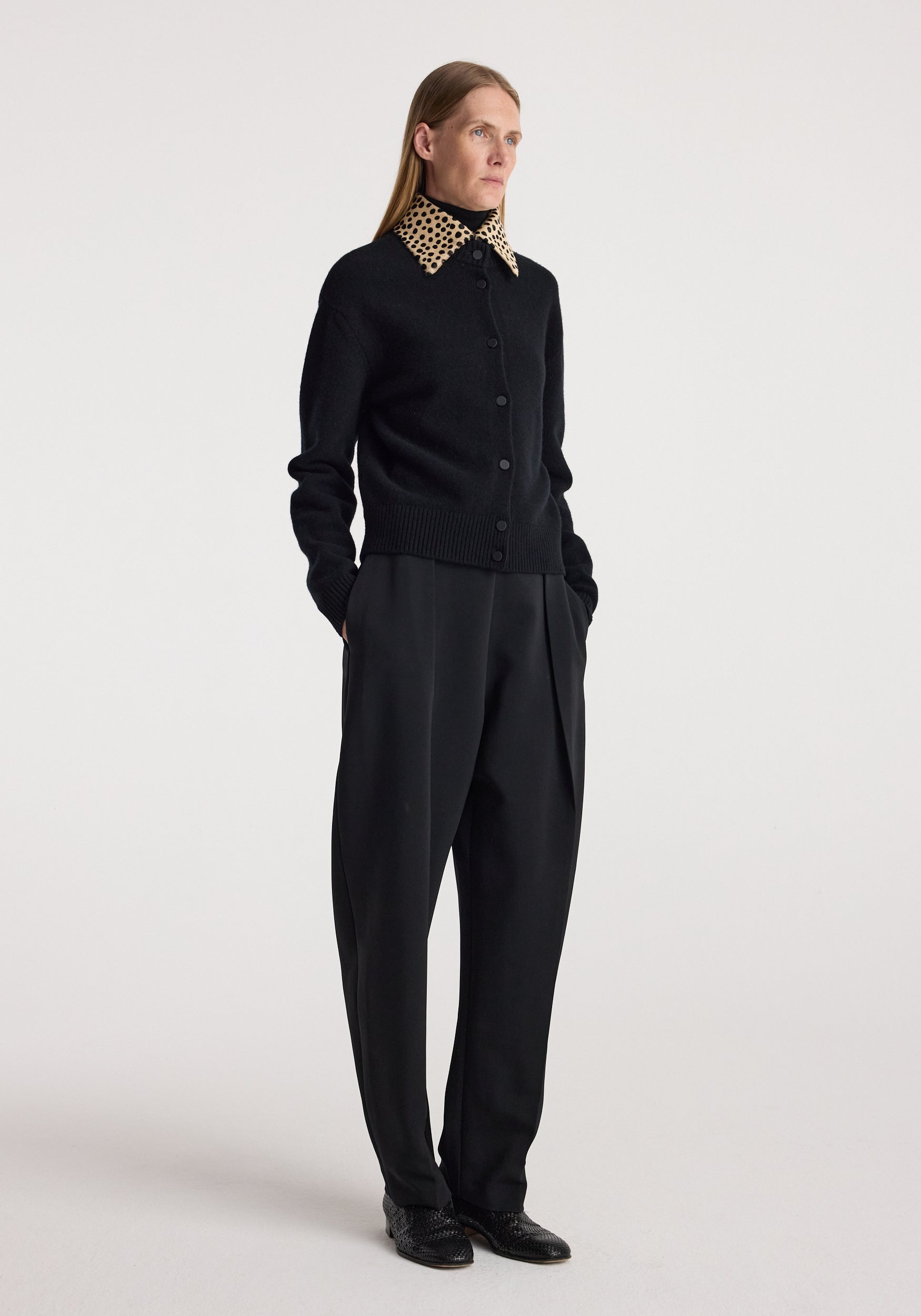 Woman wearing a black buttoned cardigan with a cheetah print collar, paired with loose black trousers and black shoes, standing against a plain background in the cheetah collar cardigan | black.