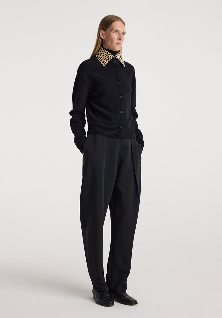 Woman wearing a black buttoned cardigan with a cheetah print collar, paired with loose black trousers and black shoes, standing against a plain background in the cheetah collar cardigan | black.