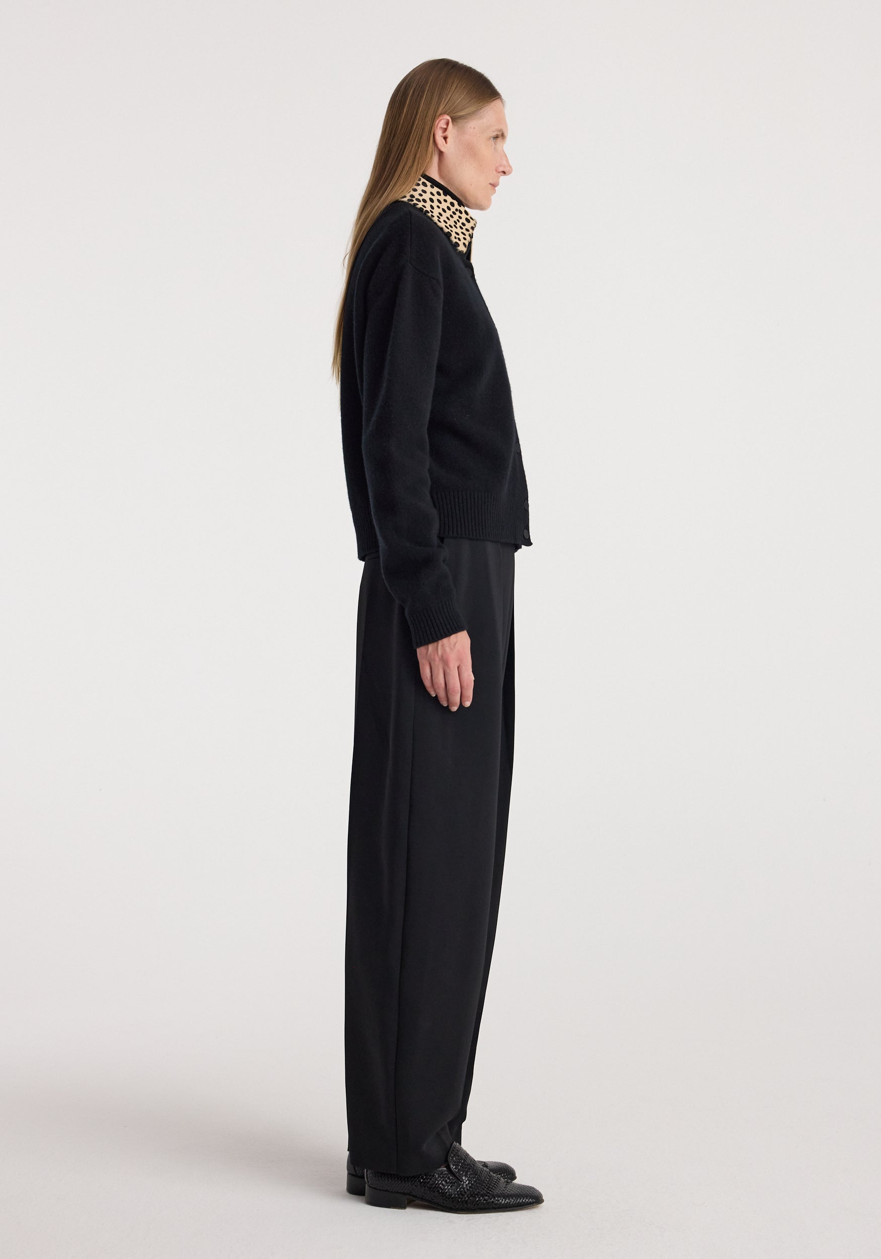 Side profile of a woman wearing a Cheetah collar cardigan | black, paired with black wide-leg pants and black shoes against a plain light background.