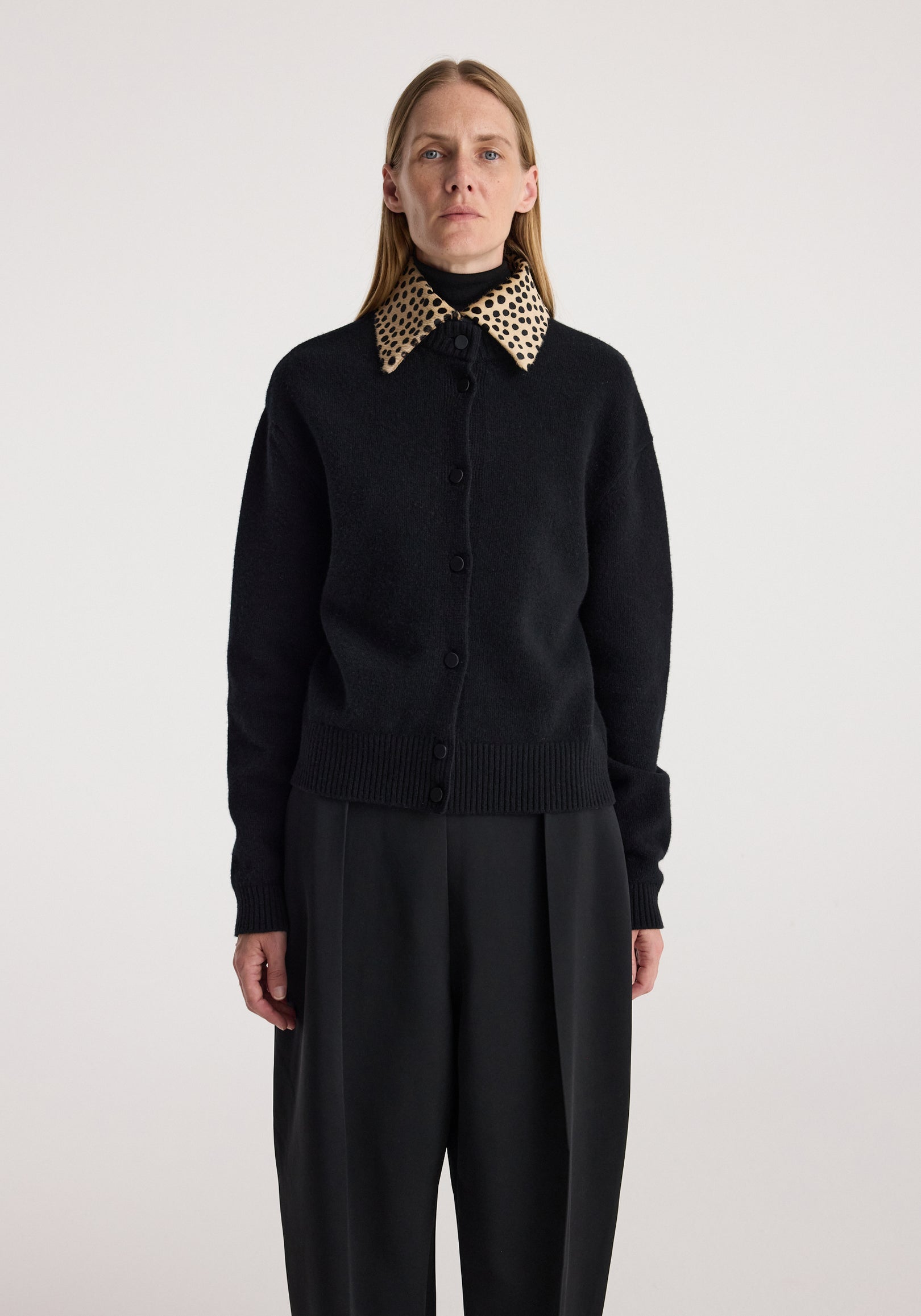 Woman wearing a black button-up cardigan with a cheetah print collar, paired with loose black pants, standing against a plain light background in the cheetah collar cardigan | black.