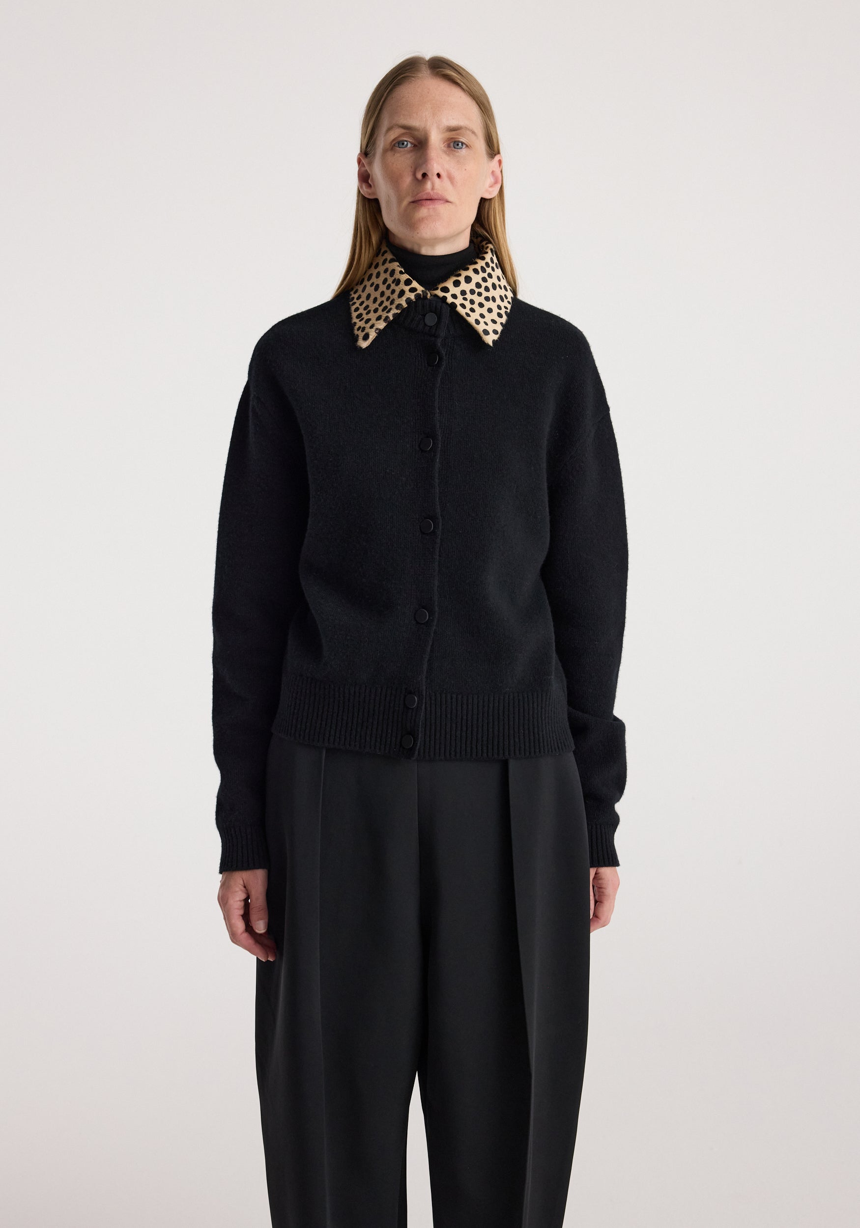 Woman wearing a black button-up cardigan with a cheetah print collar, paired with loose black pants, standing against a plain light background in the cheetah collar cardigan | black.