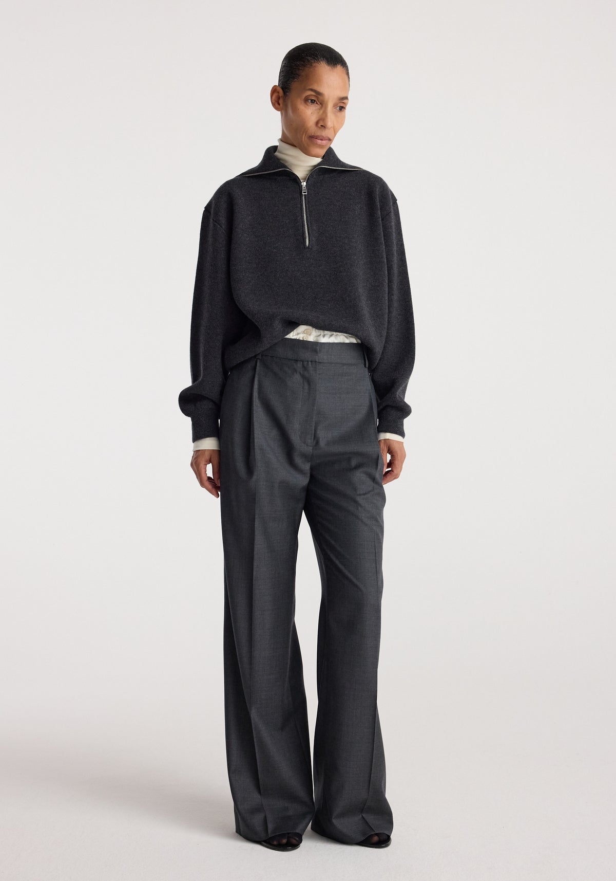 Woman wearing a charcoal Two-way zipped turtleneck | charcoal with a half-zip front, paired with loose dark trousers against a plain light background.