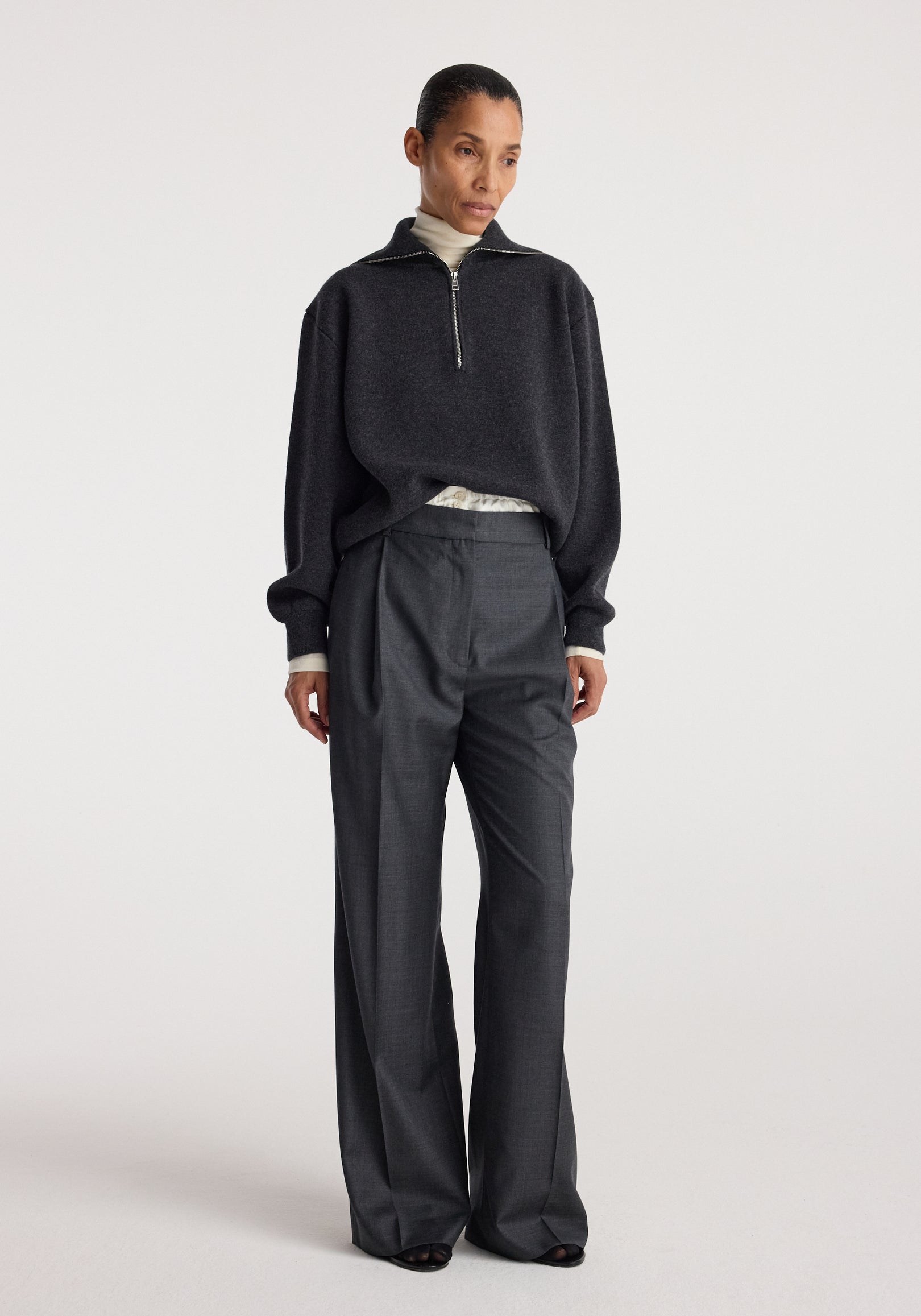 Woman wearing a charcoal Two-way zipped turtleneck | charcoal with a half-zip front, paired with loose dark trousers against a plain light background.