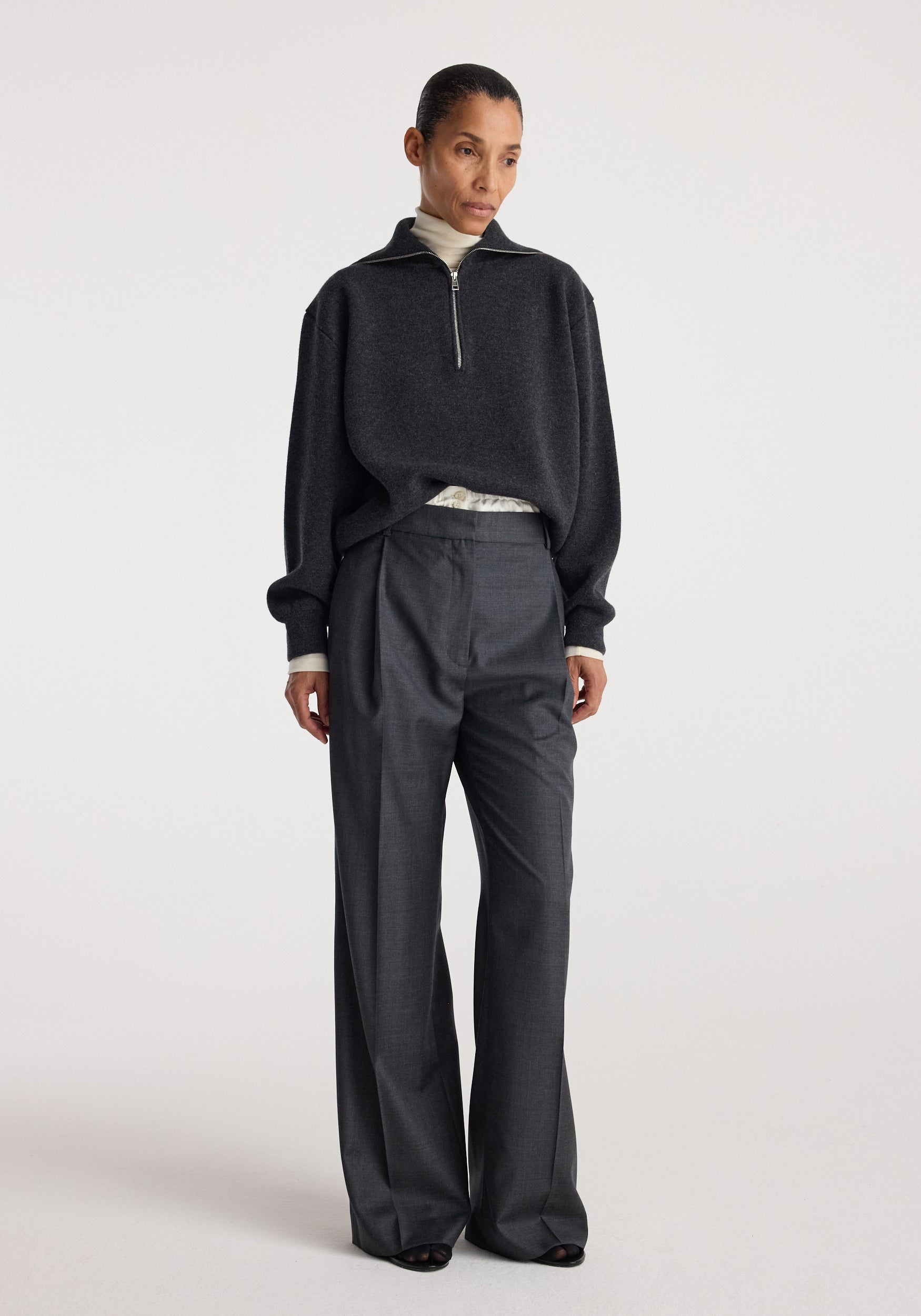 Woman wearing a charcoal Two-way zipped turtleneck | charcoal with a half-zip front, paired with loose dark trousers against a plain light background.
