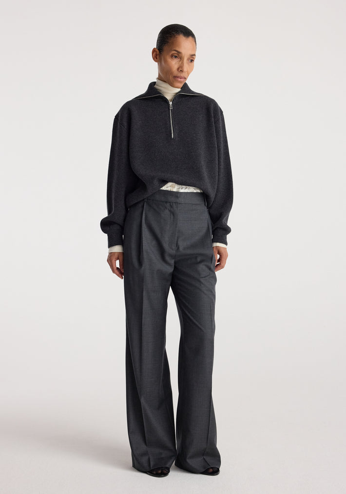 Woman wearing a charcoal Two-way zipped turtleneck | charcoal with a half-zip front, paired with loose dark trousers against a plain light background.