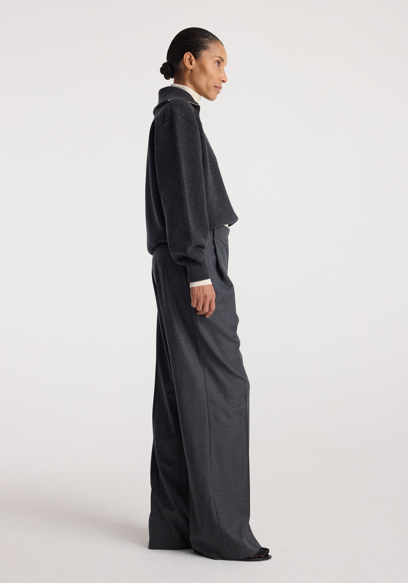 Woman wearing a charcoal Two-way zipped turtleneck | charcoal with long sleeves and loose fit, paired with high-waisted dark gray wide-leg trousers against a plain background.