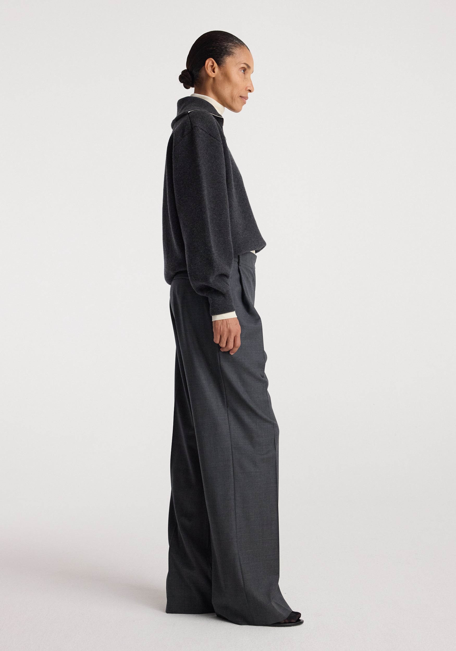 Woman wearing a charcoal Two-way zipped turtleneck | charcoal with long sleeves and loose fit, paired with high-waisted dark gray wide-leg trousers against a plain background.