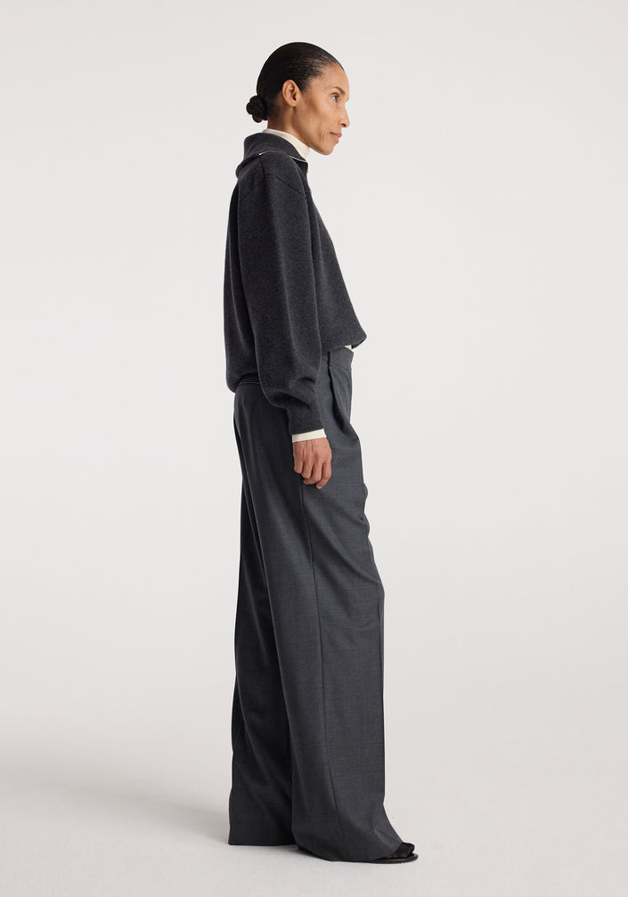 Woman wearing a charcoal Two-way zipped turtleneck | charcoal with a half-zip front, paired with loose dark trousers against a plain light background.